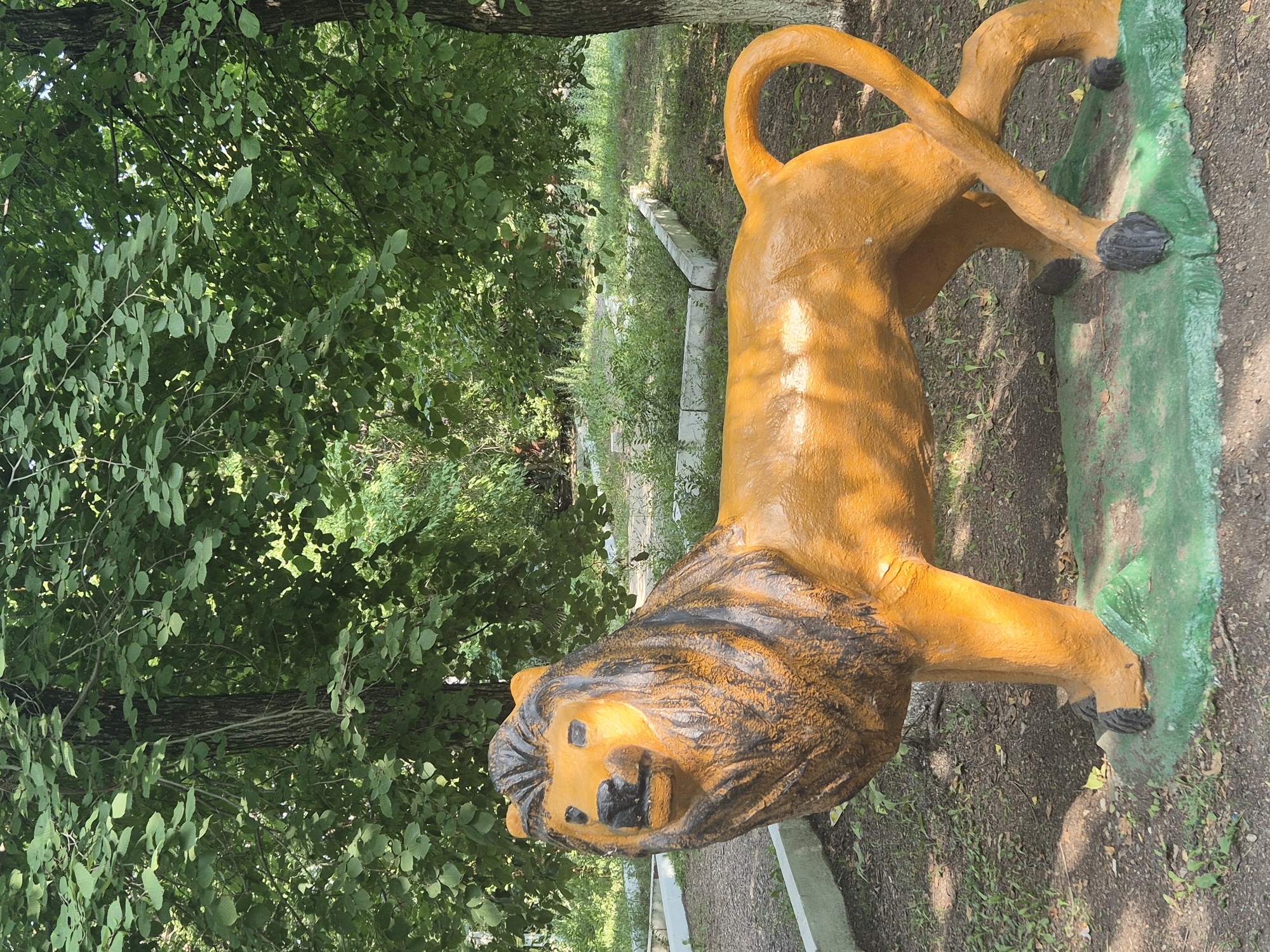 A full-body, low-angle, outdoor shot shows a weathered, orange-painted concrete statue of a lion standing on a green-painted base. The lion statue is positioned in the foreground, facing left, with its head turned slightly towards the viewer. It has a dark mane painted around its head and neck, and dark paws. The statue is located in a park-like setting with a dirt and grass ground cover, surrounded by trees with lush green foliage. In the background, there are low concrete border walls and overgrown vegetation. The scene is lit by natural daylight, casting shadows on the ground. The lion statue is likely a decorative element within the city of Новоишимское, Kazakhstan, possibly in a public park or recreational area.