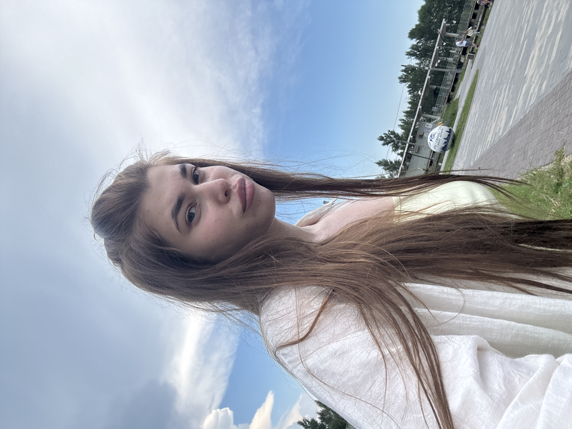 A young woman with long, light brown hair is captured in a selfie-style shot, looking directly at the camera with a slightly pursed expression. She is wearing a light yellow top and an open white linen shirt draped over her shoulders. The background suggests an outdoor setting on a partly cloudy day with a bright blue sky. In the mid-ground, a paved walkway is visible, lined with brick. Further in the background, there is a structure resembling a building or pavilion with a covered walkway, surrounded by trees. A large, decorative spherical object, possibly a globe, is placed on the ground near the structure. Several people are seen in the distance, some walking and others sitting on benches, indicating a public space. The overall impression is of a casual outdoor excursion in a park or waterfront area. While the image does not explicitly show any landmarks that pinpoint it to the city of Речицы, Russia, the presence of a public recreational area with trees and paved walkways is consistent with park environments found in many cities.