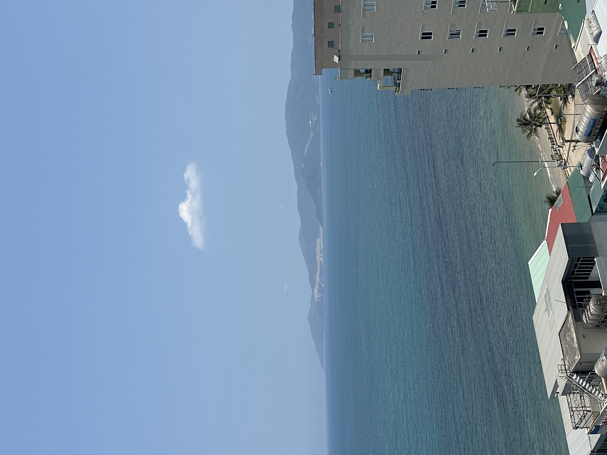 This image presents an expansive coastal scene, likely Bac Nha Trang Ward, Vietnam, viewed from an elevated perspective. The composition is dominated by a clear blue sky, a vast ocean, distant mountains, and urban development along the shore.
The top portion of the frame features a bright, light blue sky with a single, small, white cloud. Below, the calm, blue-green ocean fills the middle ground, displaying subtle ripples across its surface. In the far distance, a range of hazy, dark blue mountains or hills stretches along the horizon, blending into the atmospheric perspective. A small, white vessel, possibly a boat or ship, is faintly visible on the distant water, near the mountain range.
On the right side of the image, several multi-story buildings, likely hotels or apartments, are prominently featured. These light-colored structures have numerous windows and are built along the coastline. Their rooftops reveal various utility structures, including ventilation systems and multiple large, cylindrical metallic water tanks. One of these water tanks clearly bears the blue text "TOAN MY". A narrow strip of sandy beach or coastal area separates the buildings from the sea, adorned with several lush palm trees. A street light or utility pole stands near the beachfront.
The scene depicts a bright, sunny day, suggesting morning or early afternoon with excellent visibility. The overall atmosphere is one of tranquility and openness. There are no discernible human activities in progress. The visible Vietnamese brand name "TOAN MY" on a water tank serves as a specific location cue.