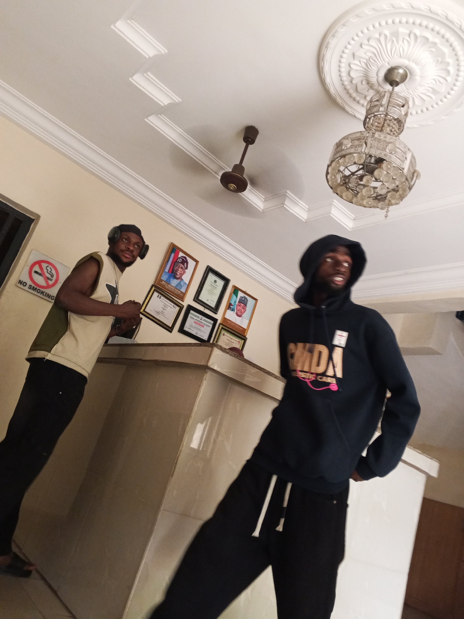 Two young men are inside a building. One is wearing a sleeveless shirt with headphones, standing near a counter. A "No Smoking" sign and framed portraits are behind him. The other man wears a hooded sweatshirt and black pants with white drawstrings. A ceiling fan and chandelier hang from the ceiling. The location is in the city of Unknown, Unknown.