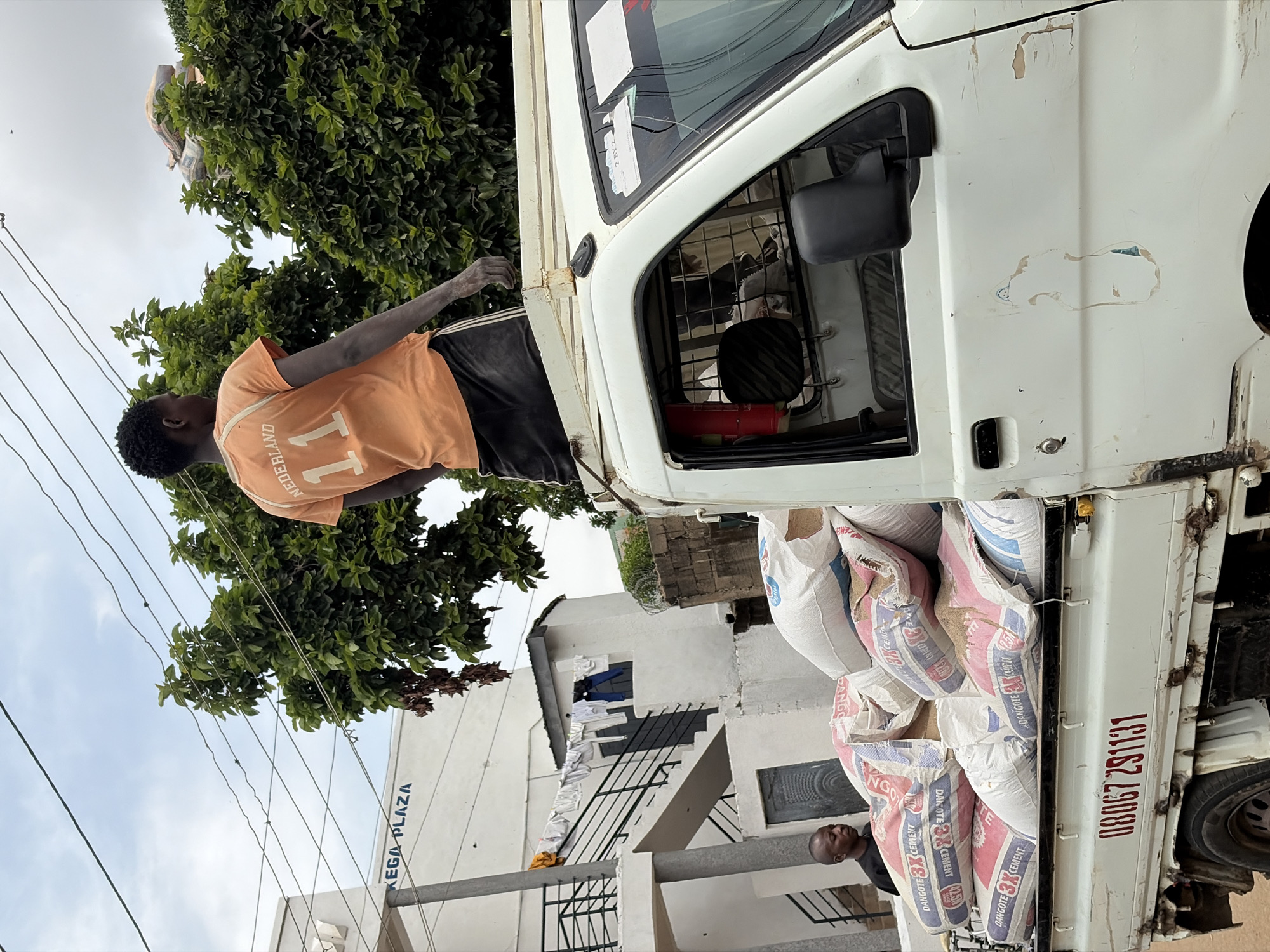 In New Stadium, Nigeria, a young man wearing an orange shirt with the number "11" and "Nederland" printed on the back stands atop a white pickup truck loaded with sacks of cement. He is looking to the right, and his hands appear dusty. Below him, inside the truck's cab, another man, wearing a dark shirt, is visible looking to the left. The truck has a phone number "08067 291131" printed on its side. In the background, a white building with "KEGA PLAZA" written on it stands near a tree-lined area. Laundry is hanging on a line on the side of a smaller white structure attached to the building. The sky is overcast.