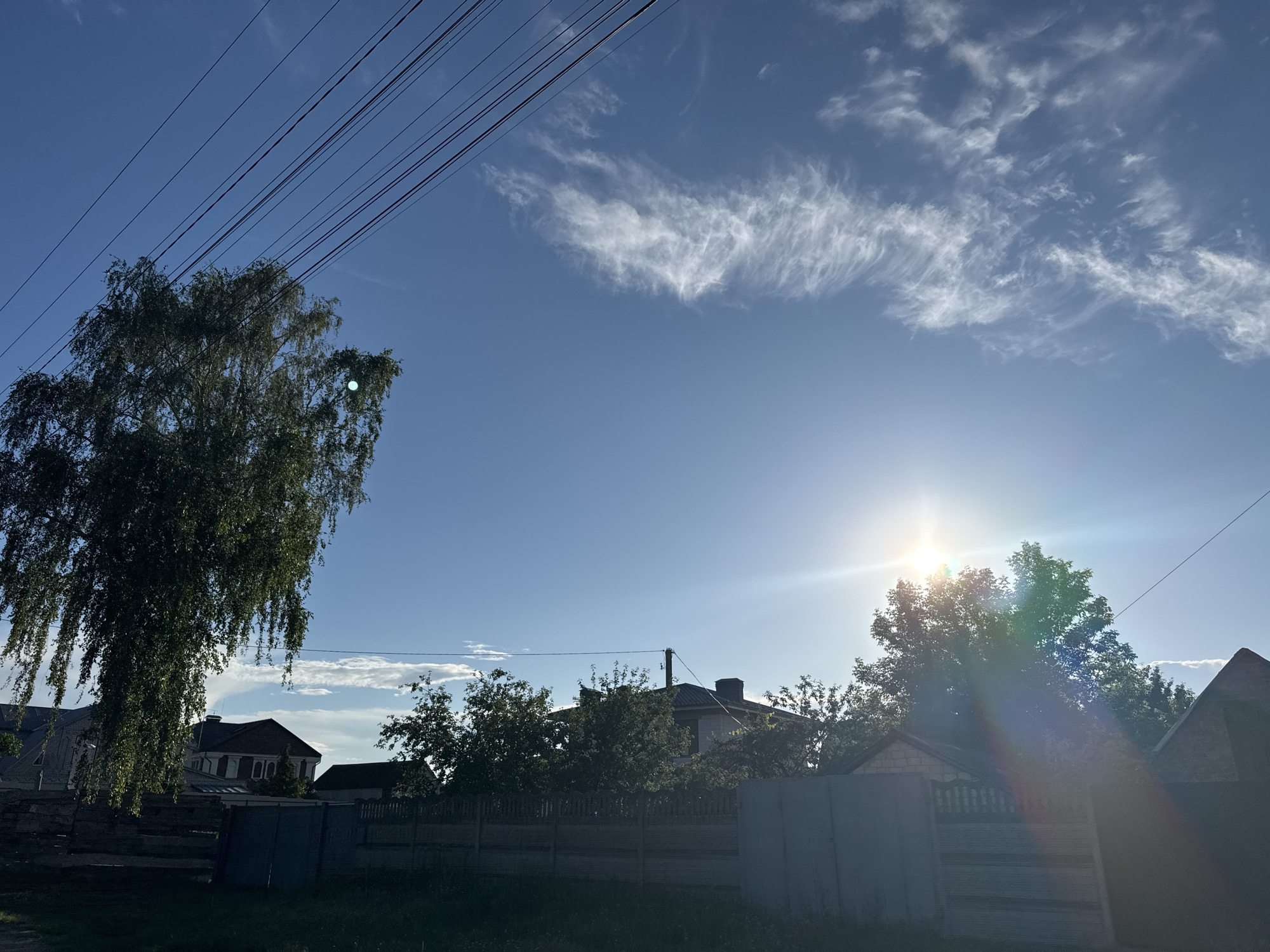 The image depicts a sunny sky with scattered clouds and power lines overhead. In the foreground, a fence runs horizontally across the frame, with a lush green tree on the left. Behind the fence, several houses with dark roofs are visible, nestled amongst more trees. The sun is a bright, direct light source in the upper right quadrant of the image, casting a lens flare that creates rainbow-like streaks across the scene. The overall environment suggests a residential area in a rural or suburban setting, likely during the daytime.