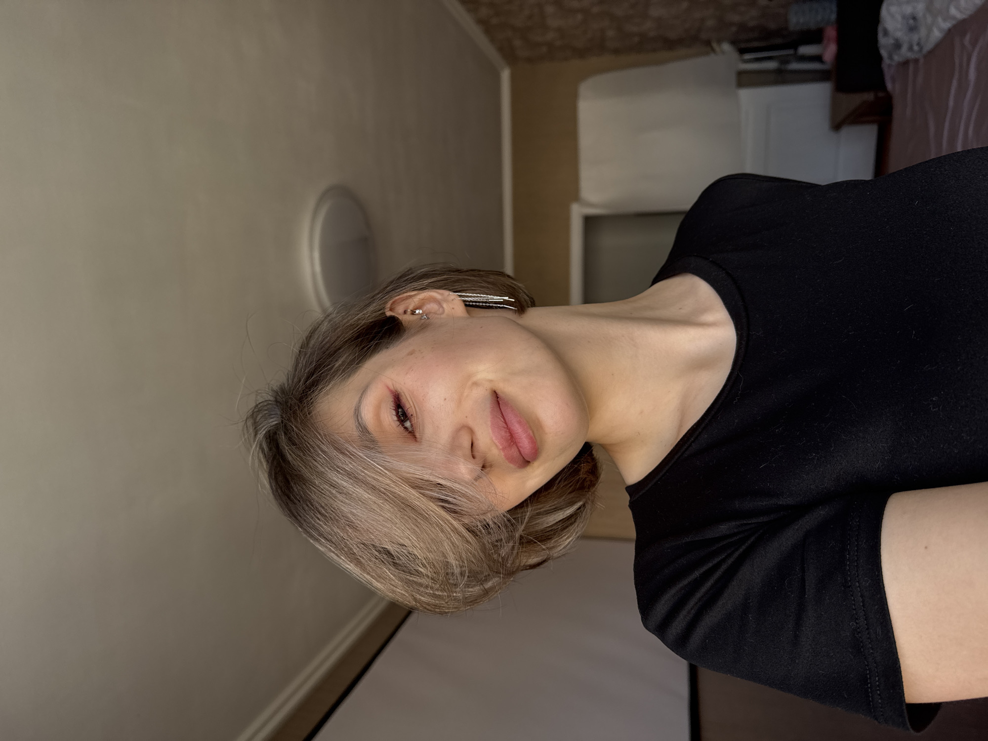 A medium shot depicts a person with short, light-brown hair and a fair complexion. They are wearing a black top. The background includes a beige wall with a circular light fixture. A blurred, undefined interior space with furniture is also visible. The location is unknown.