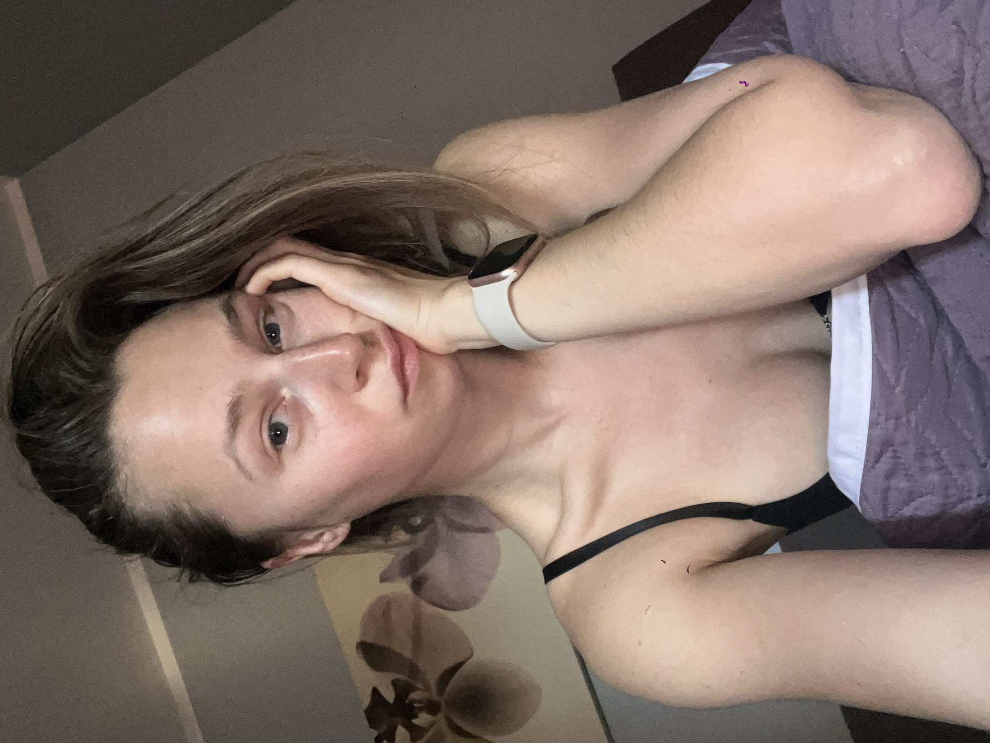 A fair-skinned woman with shoulder-length brown hair is lying on a bed with a light purple quilt. She is wearing a black bra and an Apple Watch with a white band on her left wrist. Her left hand is resting on her cheek. The background includes a wall with a floral design, and the room appears to be indoors. Bečići, Montenegro, is not discernible from the visual elements of this image.