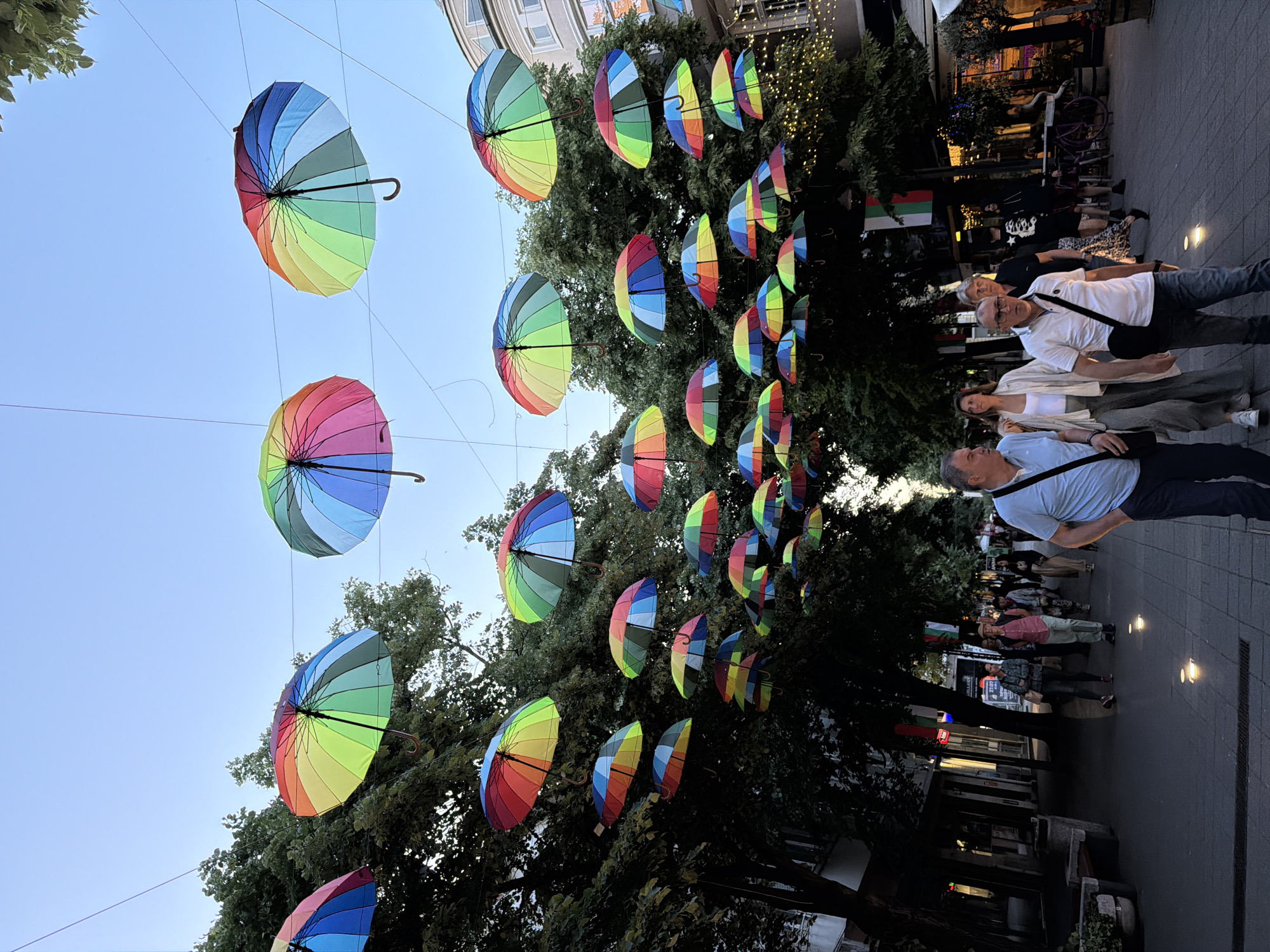 The image captures a street scene in Burgas, Bulgaria, adorned with colorful, rainbow-striped umbrellas suspended above the pedestrian walkway. The umbrellas are arranged in rows, creating a vibrant canopy overhead. Below, people stroll along the paved street, including a group of four individuals in the foreground, one of whom is wearing a blue shirt. Trees line the sides of the street, with their foliage interspersed with the hanging umbrellas. Buildings are visible in the background, along with street lamps providing lighting. The overall scene has a festive and lively atmosphere.