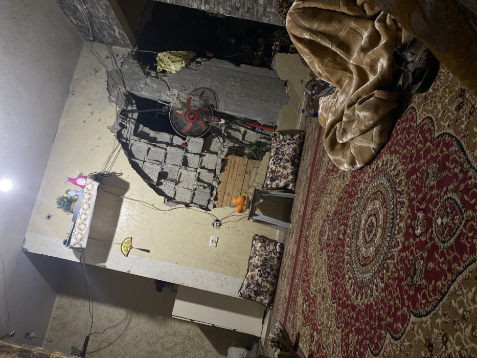 The room shows significant damage with a large hole in the wall, exposing concrete blocks. A red fan is visible within the opening. An ornate rug covers much of the floor. A shelf with decorative items is on the intact wall. Pillows sit on metal chairs near the damaged area. An orange and possible repair wood are near the damaged wall. A beige blanket and a pair of shoes rest on the rug.