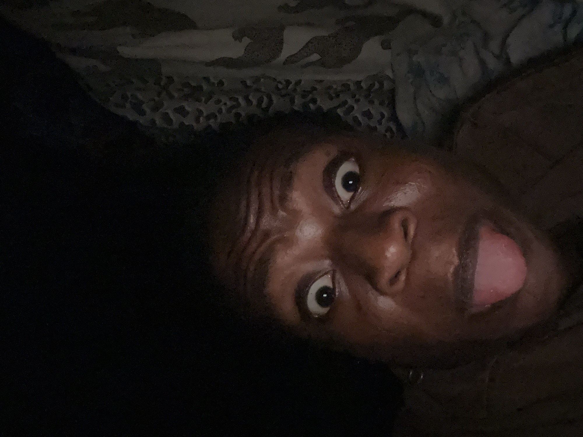 A close-up, angled view shows a person with dark skin lying down. They have wide eyes and their tongue sticking out. Their dark hair is visible to the left of their face. The background consists of a patterned blanket or sheet with what appears to be a leopard print design.