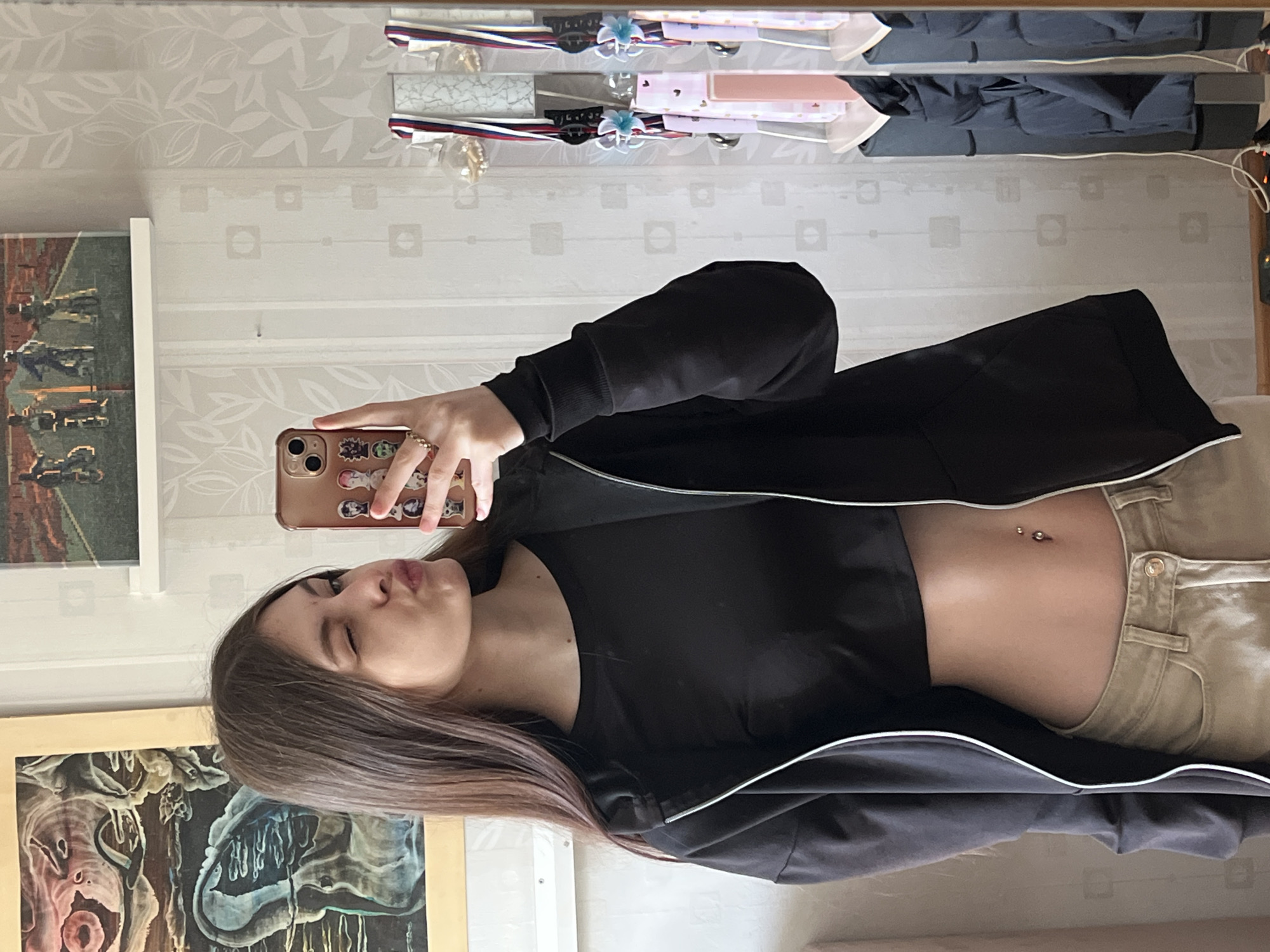A young woman with long brown hair is taking a selfie in a mirror. She is wearing a black crop top, khaki pants, and a black jacket. Her midriff is exposed and she has a belly button piercing. She is winking and puckering her lips. Her phone has a brown case with cartoon stickers.
Behind her are two framed artworks hanging on a light-colored, patterned wall. Above, a shelf or ledge is visible, holding miscellaneous items. The setting appears to be an indoor space. It cannot be determined if the location is in Saint Petersburg, Russia from this image alone.