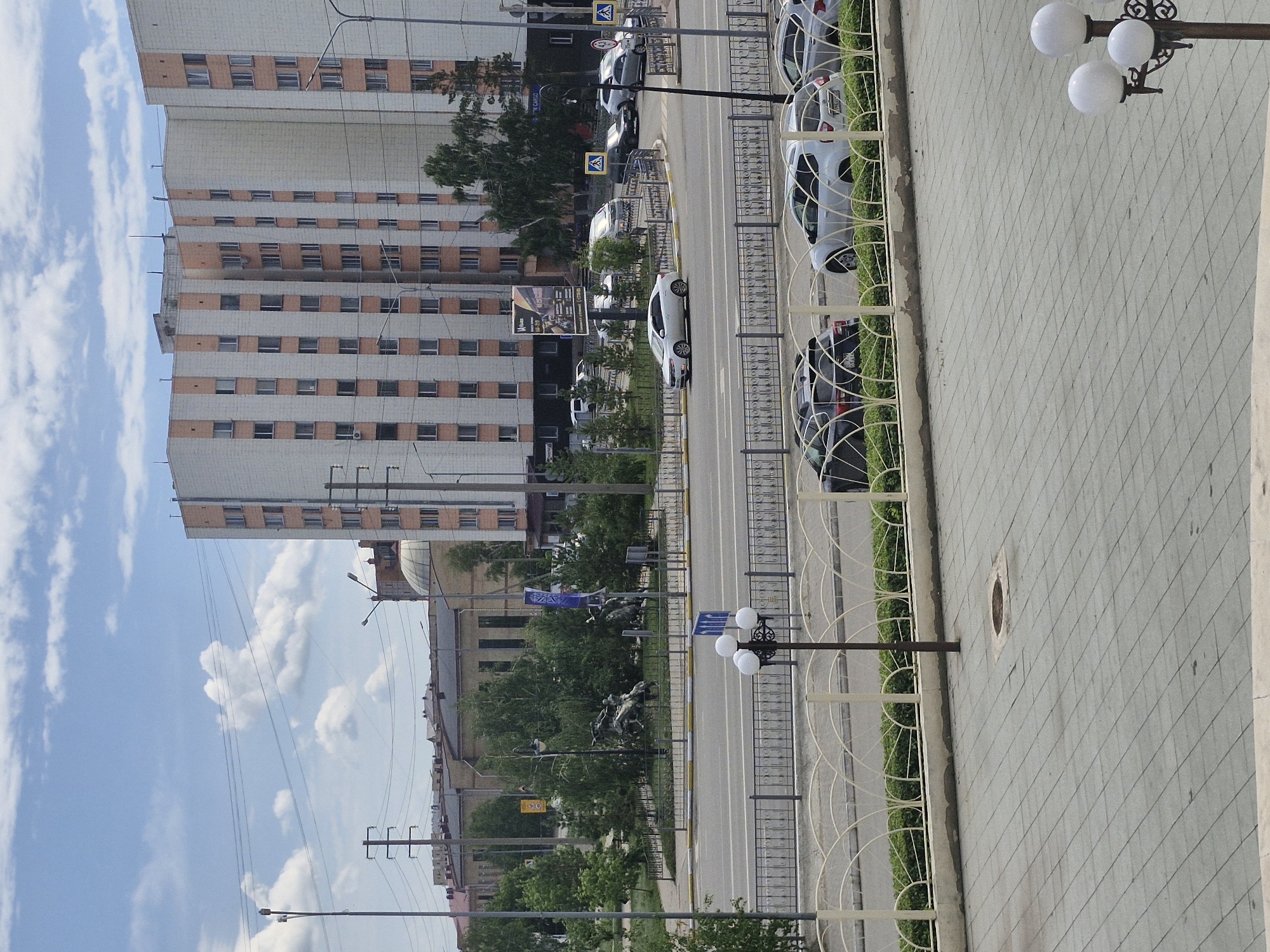 This is a medium-shot, eye-level, outdoor, daytime photograph of a street scene in Kokshetau, Kazakhstan. The background features a large, multi-story residential building with a pale facade and vertical orange accents. Power lines crisscross the sky, which is partly cloudy with cumulus clouds. In the middle ground, a street separates the viewer from the residential building. Several cars are visible on the street, including a white sedan in the left lane and a white SUV in the right lane. A circular traffic island with a statue and trees is situated at an intersection. In the foreground, a paved walkway with a decorative fence runs along the street. A street lamp with three spherical lights is present on the right side of the walkway. The overall scene depicts a typical urban environment in Kokshetau.