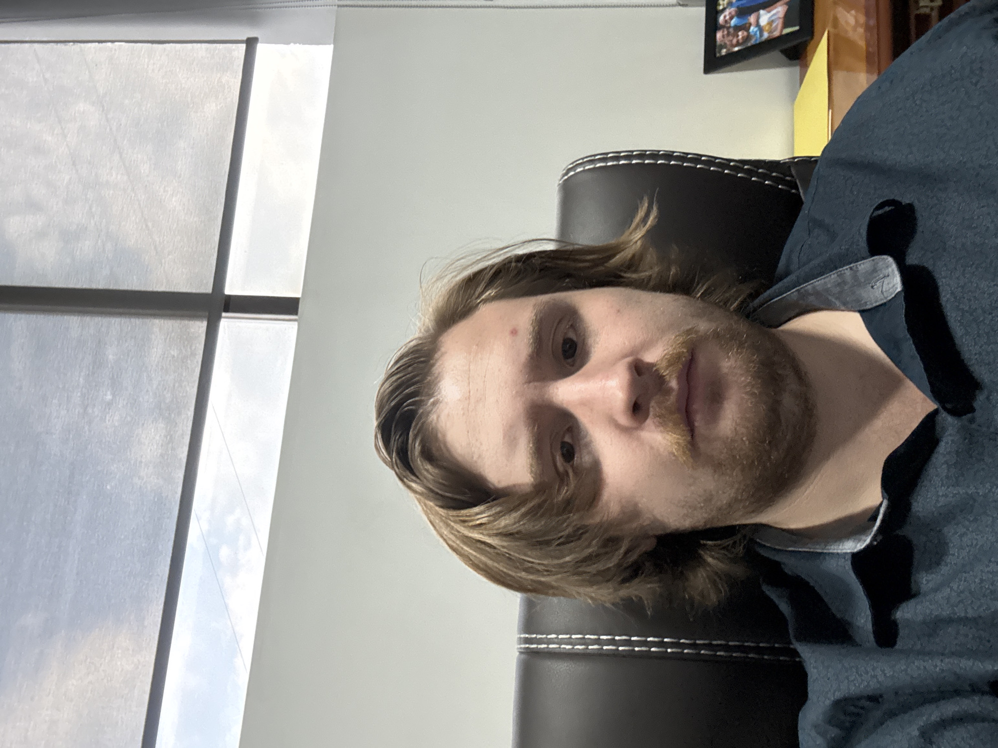 A medium shot of a person with long brown hair, a mustache, and a light beard is seated in a black leather chair with white stitching, wearing a blue button-down shirt. The scene is lit by natural light coming from a window covered with a white shade on the left side of the frame. A white wall is visible in the background, along with a framed photo and a small shelf. The location is in Cuautitlán Izcalli, Mexico. The person is looking directly at the camera.