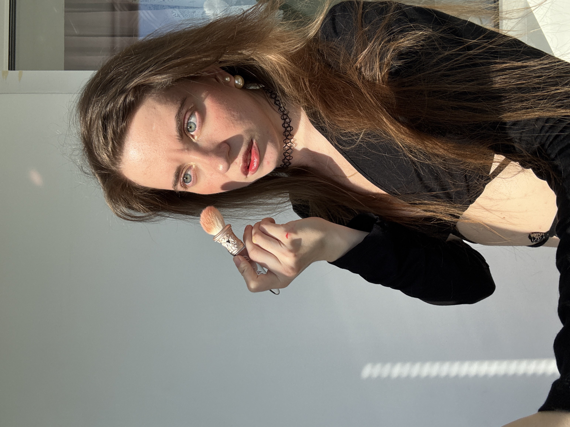 A woman with long brown hair is holding a makeup brush. She is wearing a black top and a black choker necklace. Her eyes are blue and she has makeup on her face, including lipstick. There is a pearl earring in her left ear. The image appears to be taken indoors, with natural light coming from a window, suggesting it is daytime. There is a faint shadow pattern on the wall behind her, possibly from blinds or a window frame. The background is a plain, light-colored wall and a window with a blurred reflection. The woman's expression is focused.
