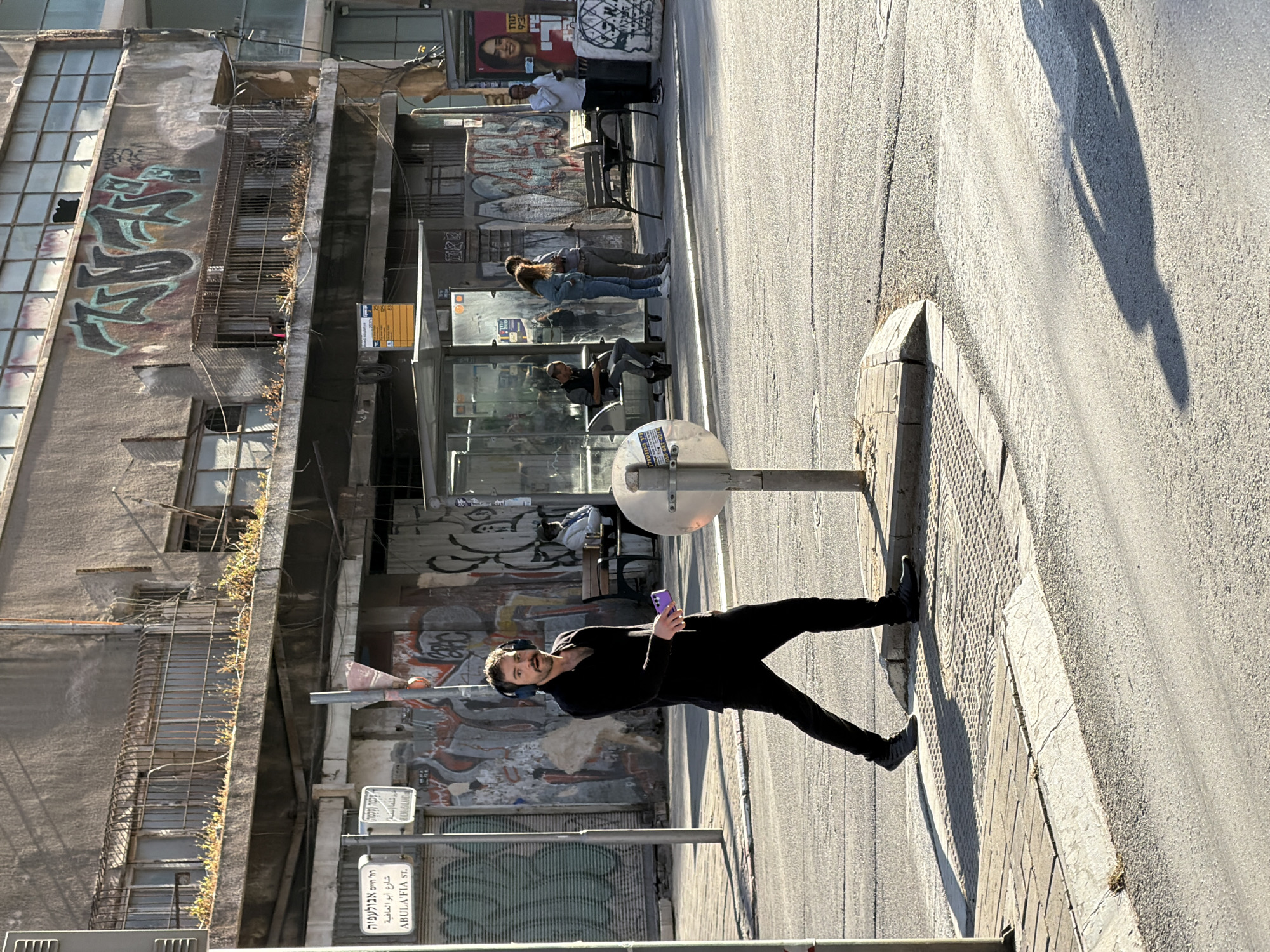 The image captures a street scene in Tel Aviv, Israel. A man in black clothing and headphones is standing on the sidewalk, holding a purple phone and facing the camera. He is positioned near a silver-colored street light fixture.
In the background, a bus stop is visible with several people waiting. A building with graffiti and deteriorating architecture lines the sidewalk, with greenery growing out of the structure. Street signs in Hebrew and English indicate that the location is Abula'fia Street. The shadow of a person or object is cast on the street.
