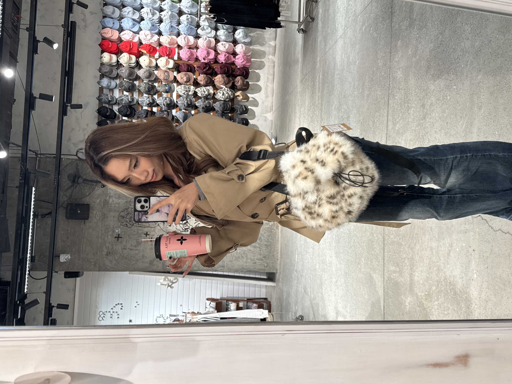 A young woman is taking a selfie in a store that appears to sell hats, as evidenced by the display of numerous caps in varying colors on the wall. She is wearing a tan trench coat and jeans, holding a pink cup, and has a patterned shoulder bag. The store has a modern, urban feel with visible ceiling fixtures and a concrete-like wall design. The location is reflected in a mirror frame, presumably within the Jianlan Road District of China.
