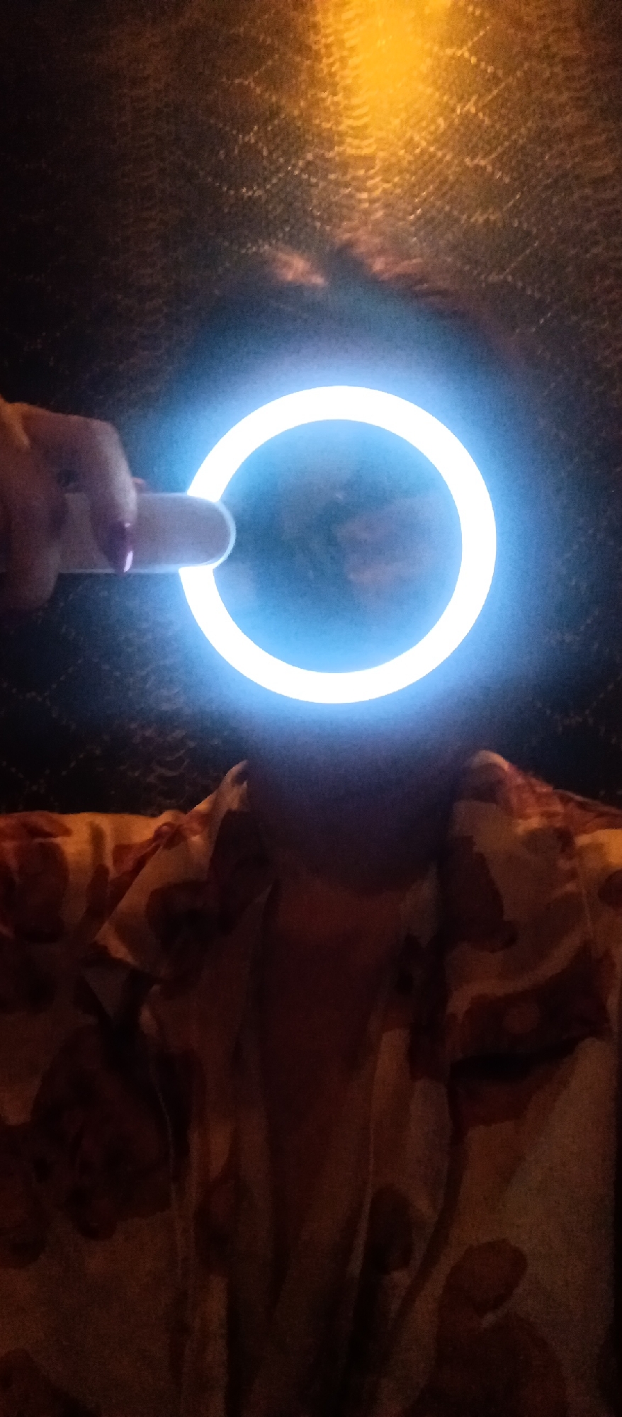 A person in an unidentifiable location, possibly indoors based on the textured, dark background, is holding a ring light in front of their face, obscuring their features. The person is wearing a patterned shirt in muted tones. A hand holding the ring light is visible on the left. The ring light emits a bright, blue-white glow.