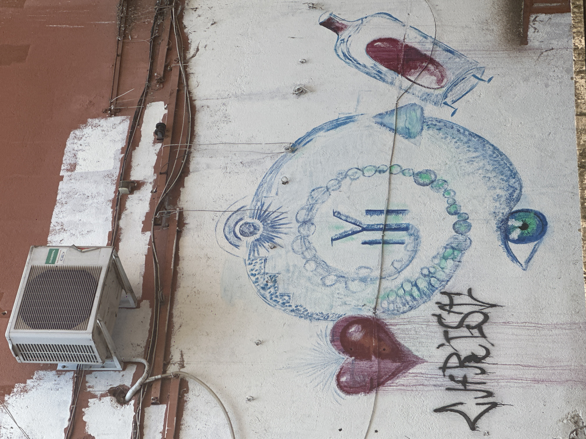 The image shows a section of an exterior wall in Pazardzhik, Bulgaria, covered in graffiti. An air conditioning unit is mounted high on the wall. Below, the wall is painted white with some sections of reddish-brown paint showing through, indicating wear. A large blue and white spiral pattern dominates the wall, with a red heart on the left and a capsule-like object on the right. The spiral is adorned with green circular shapes resembling beads. Below the heart, graffiti in black spray paint spells out "CJARISH". A green and blue eye is painted near the bottom center of the wall. Various wires and conduits run along the wall. The overall scene suggests an urban environment with visible signs of decay and artistic expression.