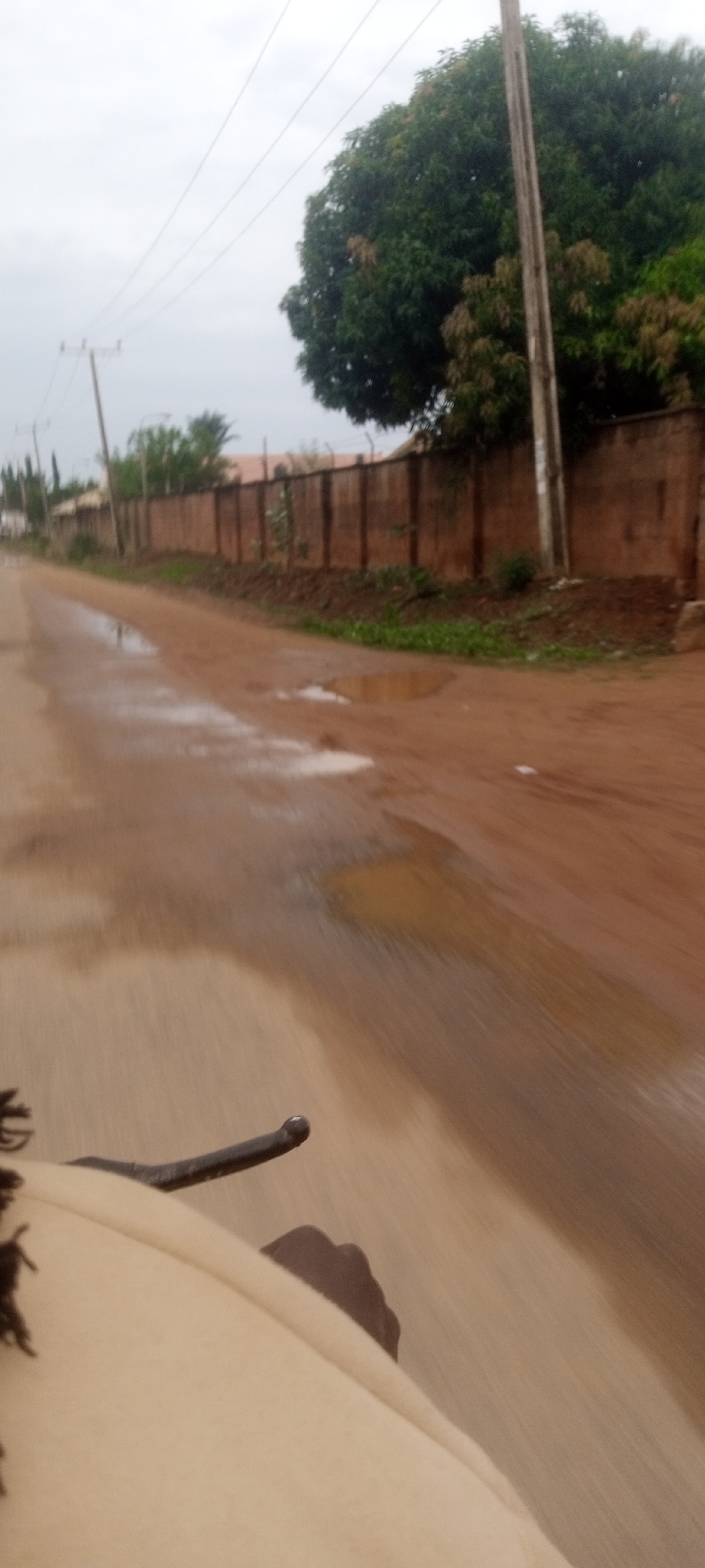 The image shows a dirt road in Narayi, Nigeria, with puddles of water on the surface. A wall runs along the right side of the road, partially obscured by a tree and power poles. On the left, there is vegetation and possibly buildings further back. The image is taken from a moving vehicle, with a hand and a handlebar visible in the foreground.