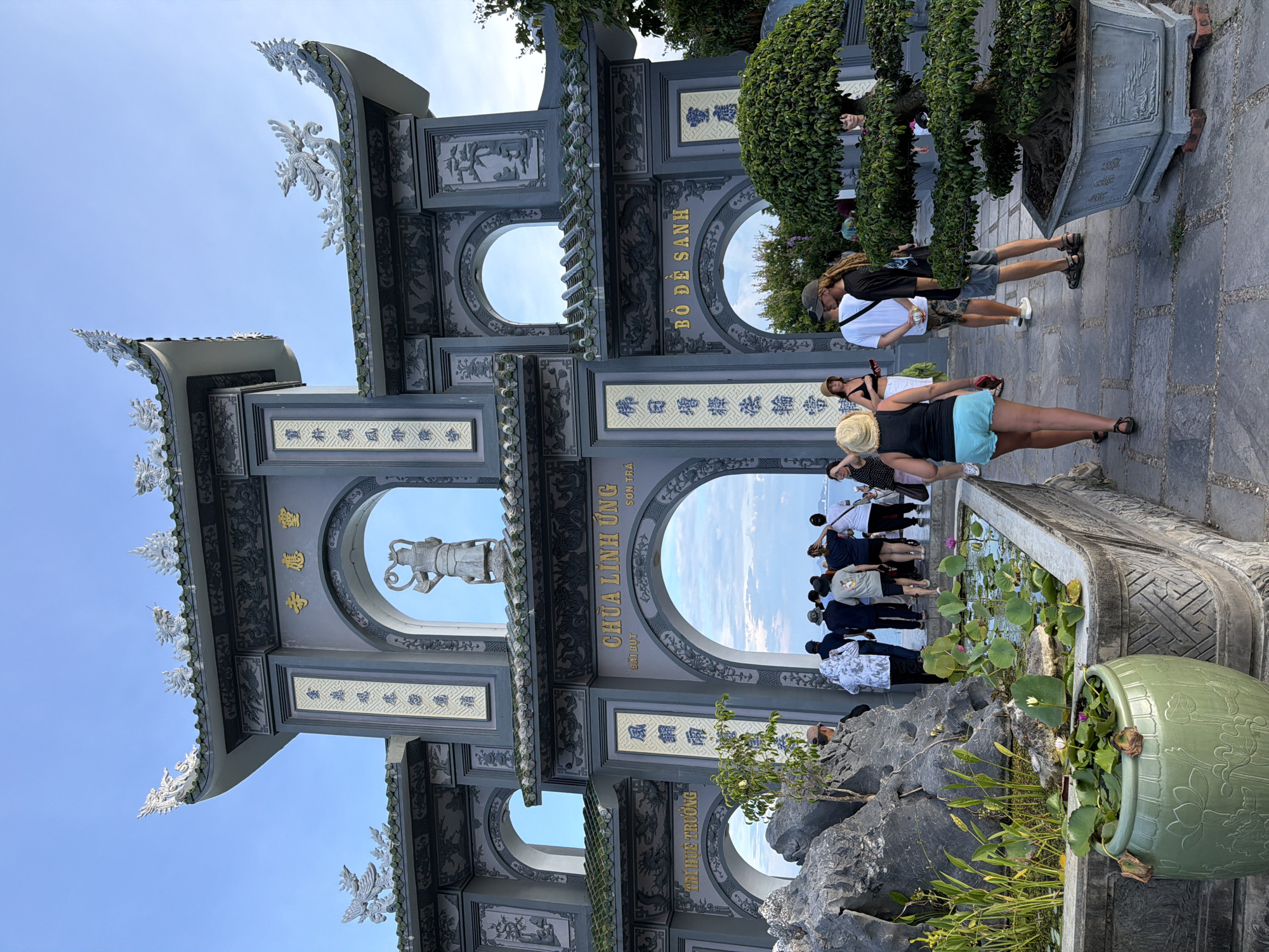 The image depicts the entrance gate of Linh Ung Pagoda, located on Sơn Trà Peninsula in Đà Nẵng, Vietnam. The pagoda's architecture is characterized by ornate, gray structures with green tiled roofs and intricate dragon sculptures adorning the eaves. The gate features multiple arched openings, with one arch revealing a statue of a deity.
A group of tourists, dressed in casual attire, are seen walking towards and standing near the gate. Among them, a woman in the foreground is walking away from the camera, wearing a black tank top and light blue shorts. To her right, a man with long dreadlocks and a woman in a straw hat are engaged in conversation. Other individuals are dispersed around the entrance.
In the lower left of the frame, a stone pond filled with lotus plants is visible, with a large green ceramic pot holding more aquatic plants placed next to it. To the right, a meticulously pruned bonsai tree in a stone planter adds to the garden-like aesthetic of the pagoda grounds. The sky is clear blue, suggesting daytime, and the overall environment appears to be a tourist attraction with cultural and religious significance.