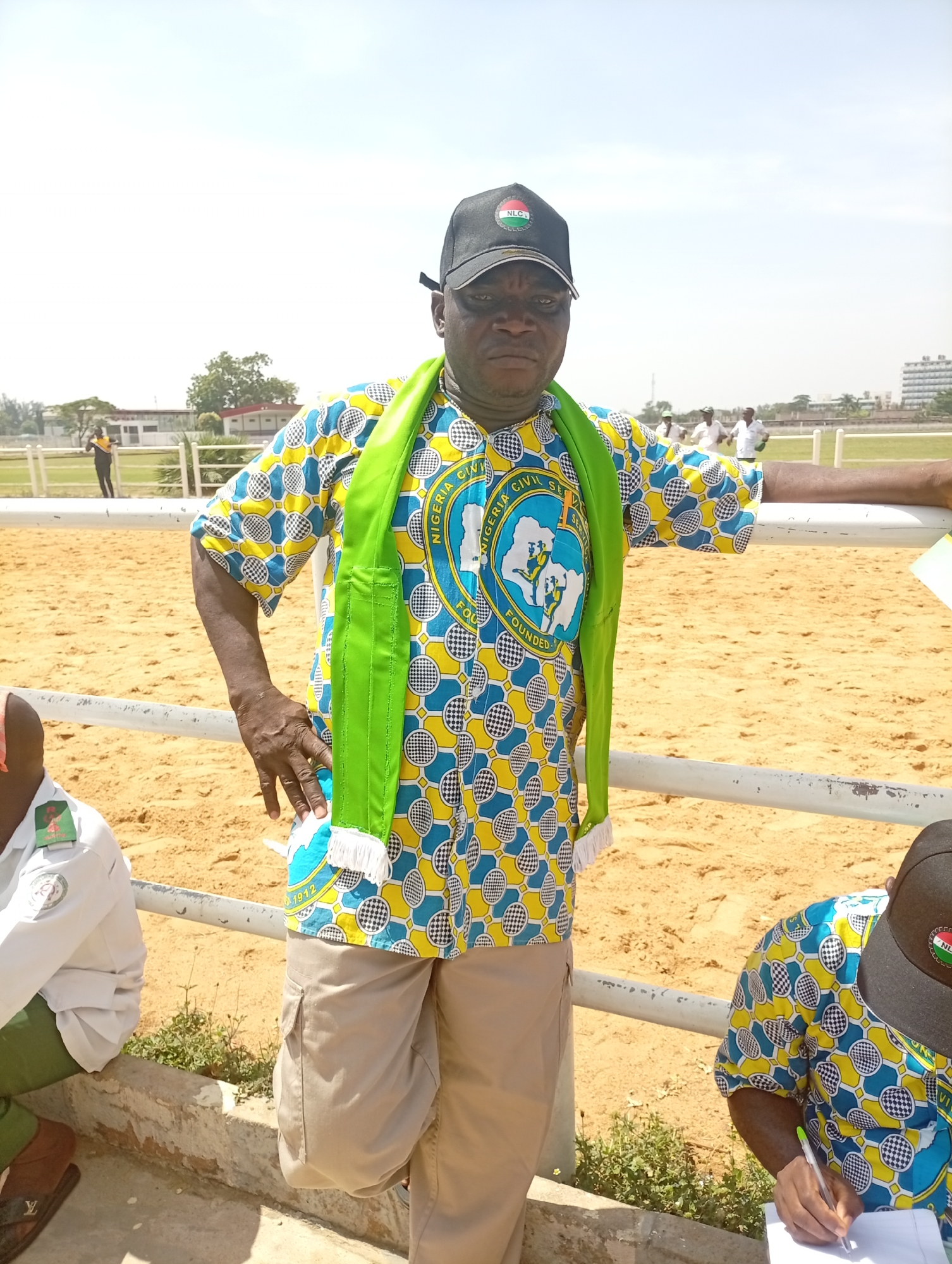 A medium shot shows a man standing next to a fence in an outdoor area. He is wearing a black baseball cap with a logo, a patterned shirt of blue, yellow, and black with a round emblem on the front, a green scarf with white fringe, and tan cargo pants. He is leaning on the fence with his right arm. The area behind the fence is a sandy or dirt surface with a building and some trees in the background. Other people are faintly visible in the distance. Two other people are partially visible in the foreground, with one wearing a matching shirt to the man standing.
Based on the logo on the man's cap and shirt, the scene is likely taking place in Nigeria. The round emblem on the man's shirt is a logo for "Nigeria Civil Service," which has the founding date of 1912. It is difficult to confirm the specific location within Unguwar Sarki, Nigeria, from the image alone.