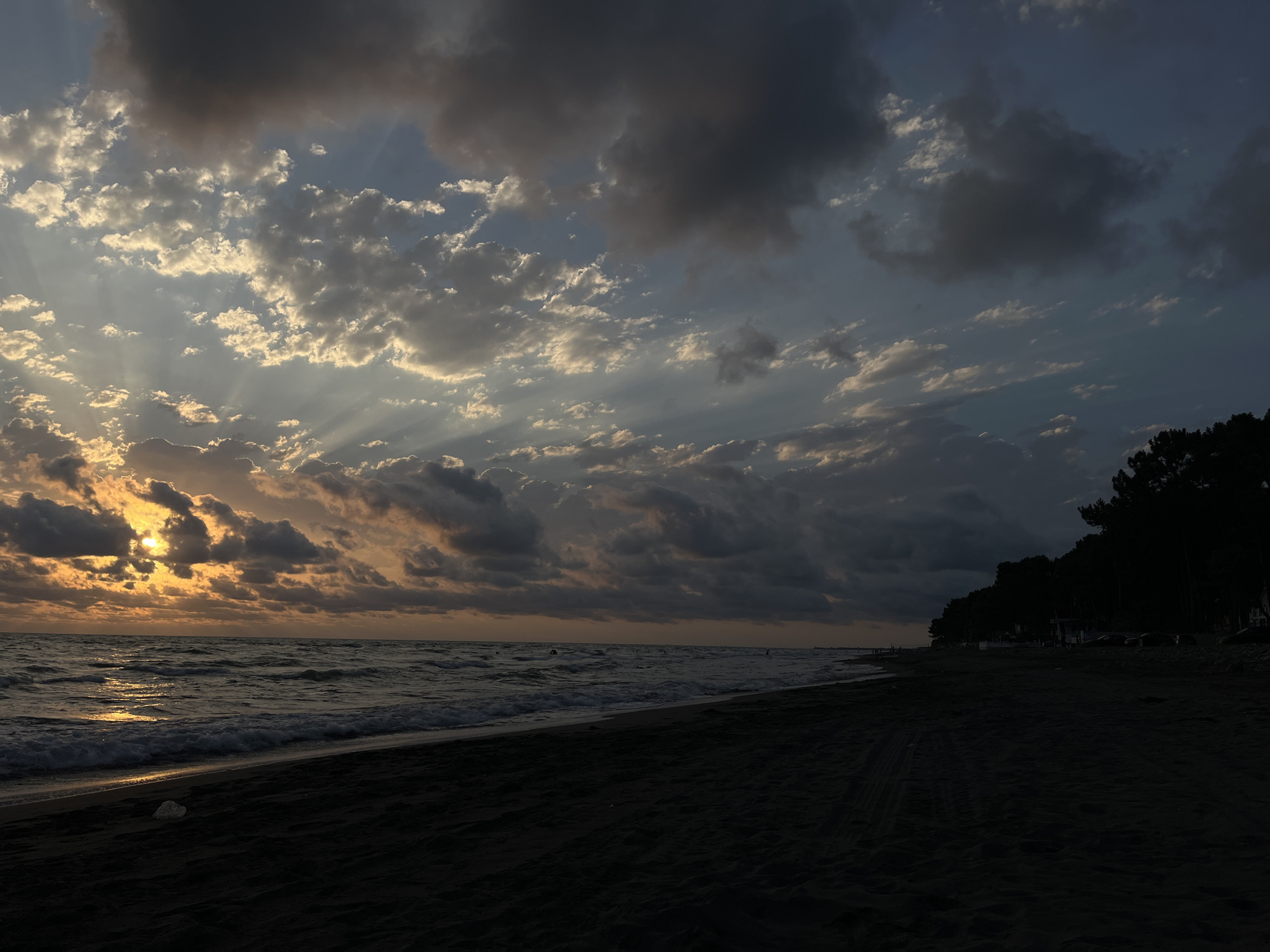 The image depicts a sunset at a beach in the village of Supsa, Georgia, located within the Supsa municipality. The scene is characterized by a dark, sandy beach in the foreground, with the gentle waves of the sea lapping at the shore. The sky is filled with dramatic clouds, with rays of sunlight piercing through them, creating a striking contrast between light and shadow. A line of trees forms a dark silhouette along the right side of the beach, suggesting a forested area adjacent to the coast. The overall atmosphere is serene and contemplative, typical of a sunset on a coastal landscape.