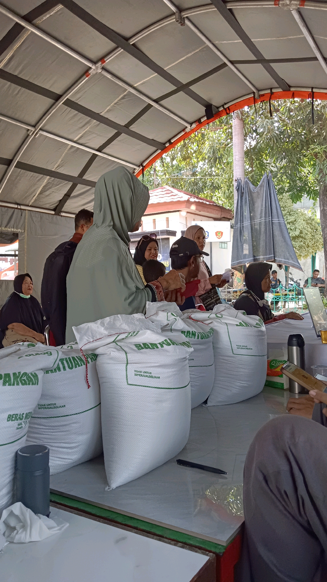 The image depicts an outdoor distribution event, likely a food aid program, under a temporary tent structure in East Jakarta, Indonesia. Several people are gathered, receiving or distributing sacks of rice. The sacks are white with green lettering, some of which is legible as "BANTUAN PANGAN" (Food Aid) and "BERAS MEDIUM" (Medium Rice).
In the foreground, a person in a grey hijab is interacting with a table displaying the rice sacks. To their left, another person in a black hijab is seated. Behind them, several other individuals, including men and women, are visible, some wearing headscarves. One woman in a yellow shirt and pink headscarf is smiling.
A table runs along the front of the scene, laden with the rice sacks. On the table, a silver thermos, a pen, and some papers are present. In the background, beyond the tent, trees and a building with red-tiled roofs are visible, suggesting a semi-urban or suburban setting. The presence of a motorcycle in the far background further indicates the location. The overall atmosphere appears to be one of organized distribution and community gathering.