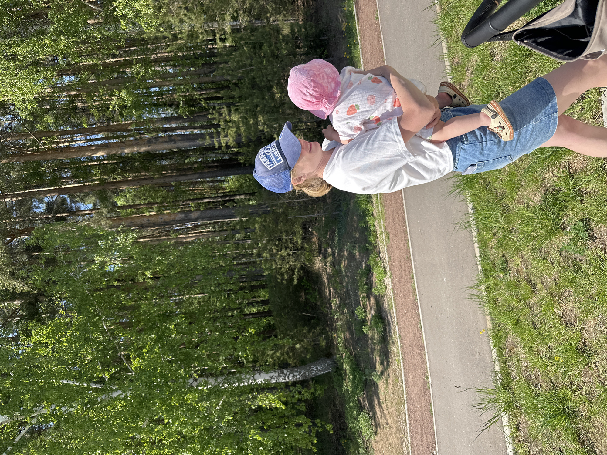 A woman in a white t-shirt and blue denim shorts holds a child wearing a pink sun hat and a white shirt with red strawberry prints. The woman wears a blue baseball cap. They stand near a paved path in a grassy area bordered by a forest. A stroller is partially visible in the top right corner. The location is in the city of Chaikovsky, Russia.