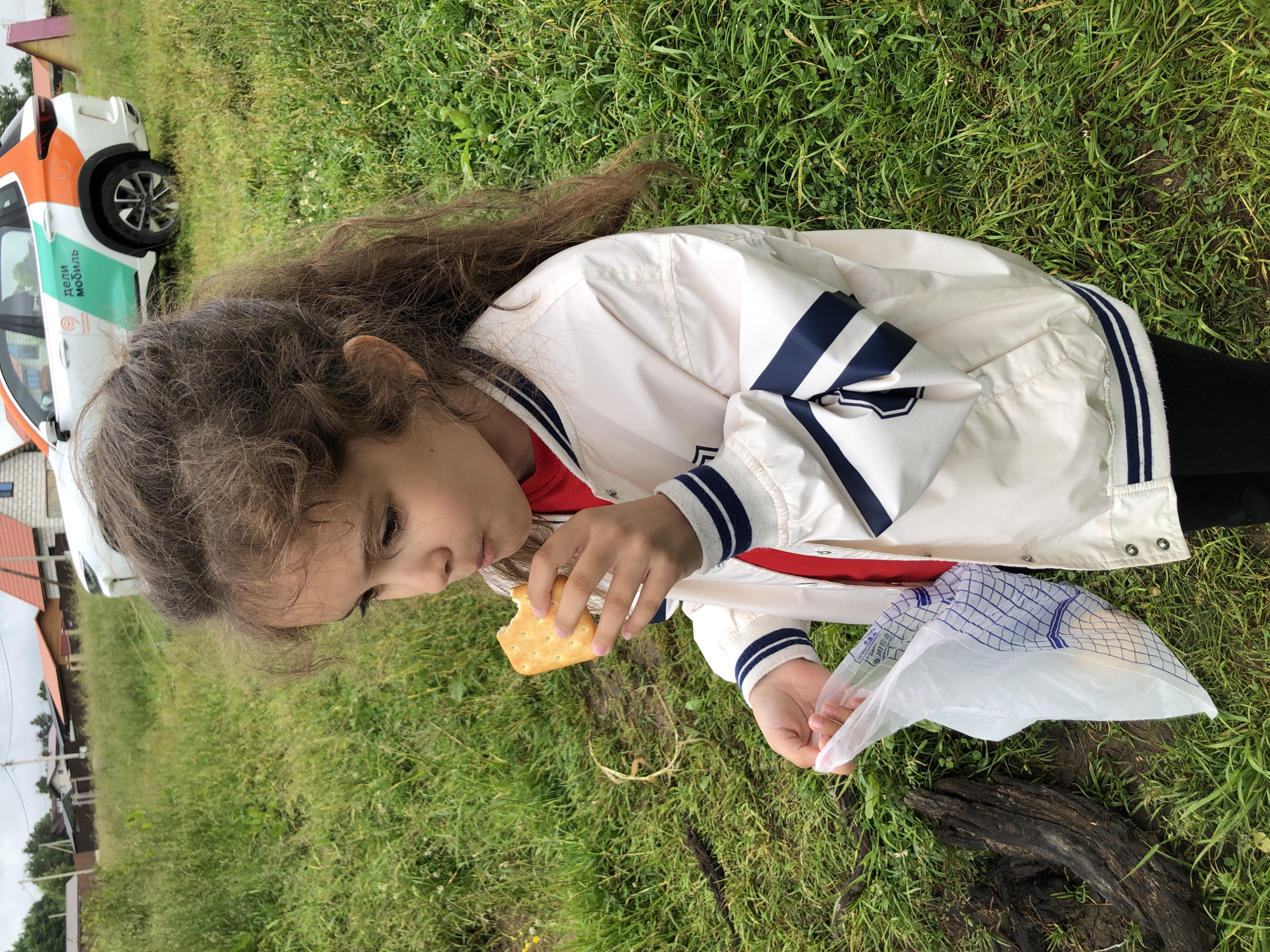 A young girl with long, dark, wavy hair is sitting on the grass, eating a cracker. She is wearing a white jacket with navy blue stripes on the sleeves and collar, and a red shirt underneath. Her fingernails are painted pink. In her left hand, she holds the cracker, and in her right hand, she holds a white plastic bag with a blue grid pattern.
The setting is outdoors, with lush green grass and some scattered clover and small white flowers. A dark, weathered piece of wood lies on the ground in the foreground. In the background, a white car with orange and green accents is parked. The car has text on its side that appears to be in Russian, with the word "МОБИЛЬ" visible, suggesting it might be a service vehicle. Further in the background, houses with red roofs are visible, indicating a rural or suburban setting.
The lighting suggests it is daytime, possibly overcast, as there are no strong shadows. The overall mood seems casual and relaxed, as the girl is enjoying a snack outdoors. The crops highlight the girl's face and hands, the details of her clothing, the cracker, the plastic bag, and the surrounding grass and wood. The presence of the car and houses provides context for the location.