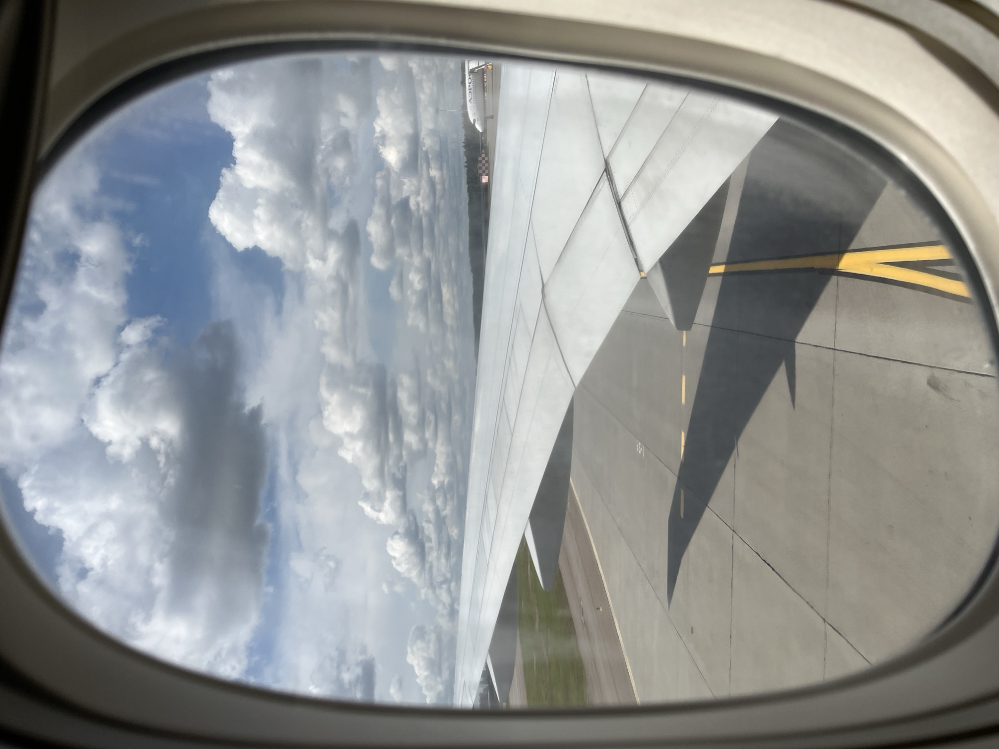 The image is taken from an airplane window, looking out at a runway. A large airplane wing dominates the foreground, casting a shadow on the tarmac. In the distance, another airplane is visible, with the word "A320" printed on its side. The sky is filled with white, fluffy clouds against a blue background. The scene appears to be at an airport, possibly on the outskirts of the city of Rusanovo, Russia, given the presence of an Aeroflot airplane. The overall impression is one of travel and anticipation.