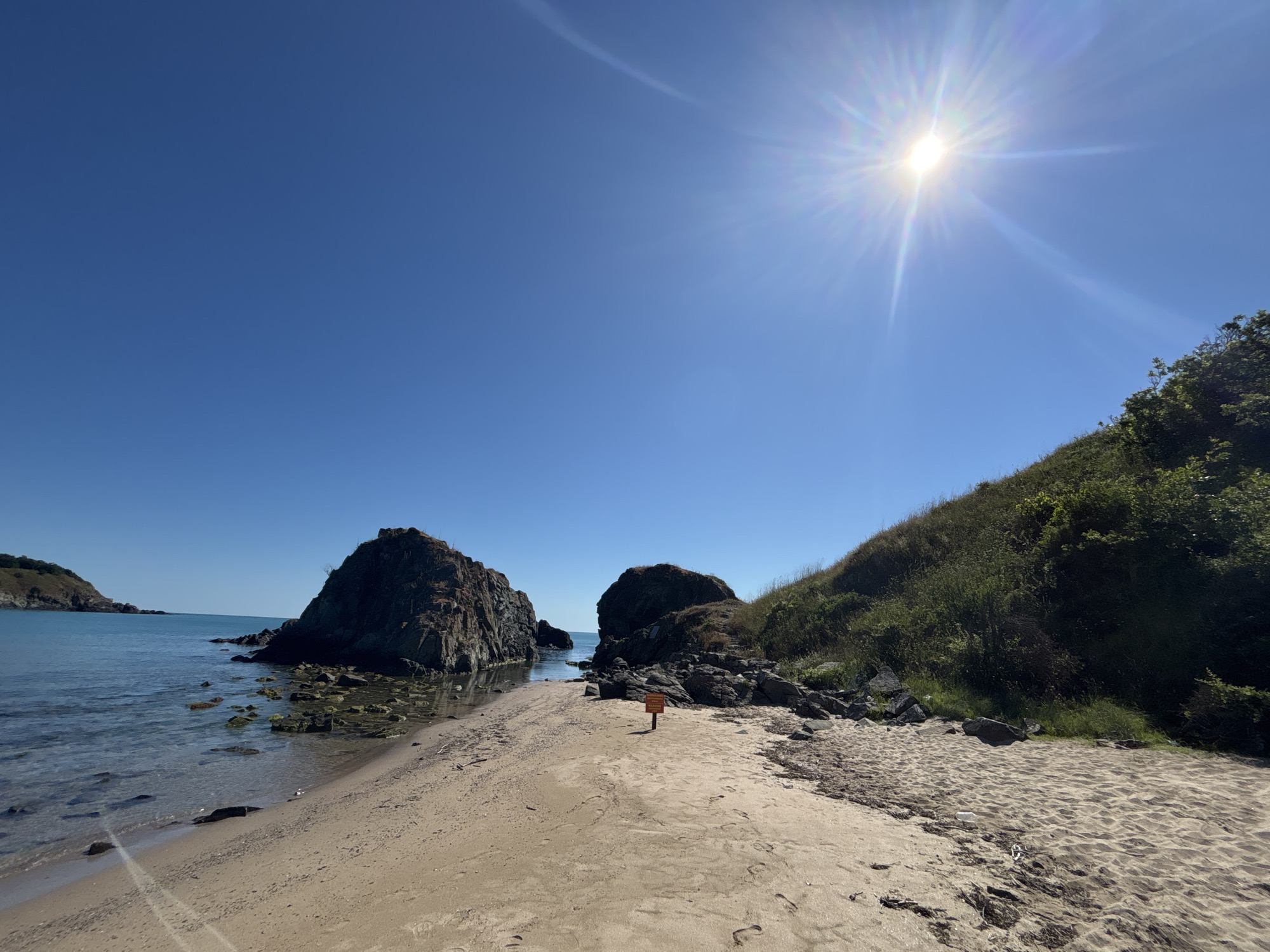 This is a wide-angle, eye-level, outdoor, daytime photograph of a beach scene with two large rock formations in the sea, taken on a clear, sunny day. The sun is high in the sky, casting a bright glare and lens flare. The water is calm and transparent, showing rocks beneath the surface near the shore. The beach is sandy with some scattered seaweed and footprints. In the middle of the beach, a small orange sign on a pole is visible. A lush green hillside rises on the right side of the image, extending into the background. The left side shows the sea meeting another small, green, hilly landmass in the distance. The scene depicts a natural coastal environment with clear blue skies. The location is identified as the beach in Rezovo, Bulgaria.