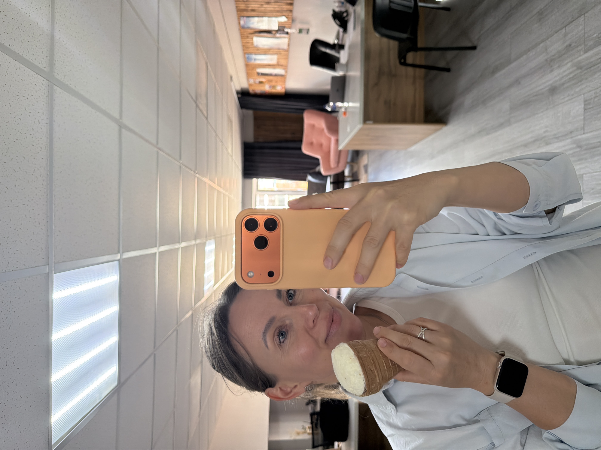 A woman is taking a selfie in an office setting. She is holding a white and brown object resembling a root vegetable in one hand and a peach-colored smartphone in the other. The woman is wearing a white t-shirt, a light blue button-down shirt, and a silver smartwatch. She also has a silver ring on her finger. The office has tiled ceilings with recessed fluorescent lights, a light wood floor, and black chairs. In the background, there is a glimpse of a pink chair and a wooden desk. Given the context of the request for the city of Chaikovsky, Russia, it can be inferred that the photo was taken in an office located in this city.