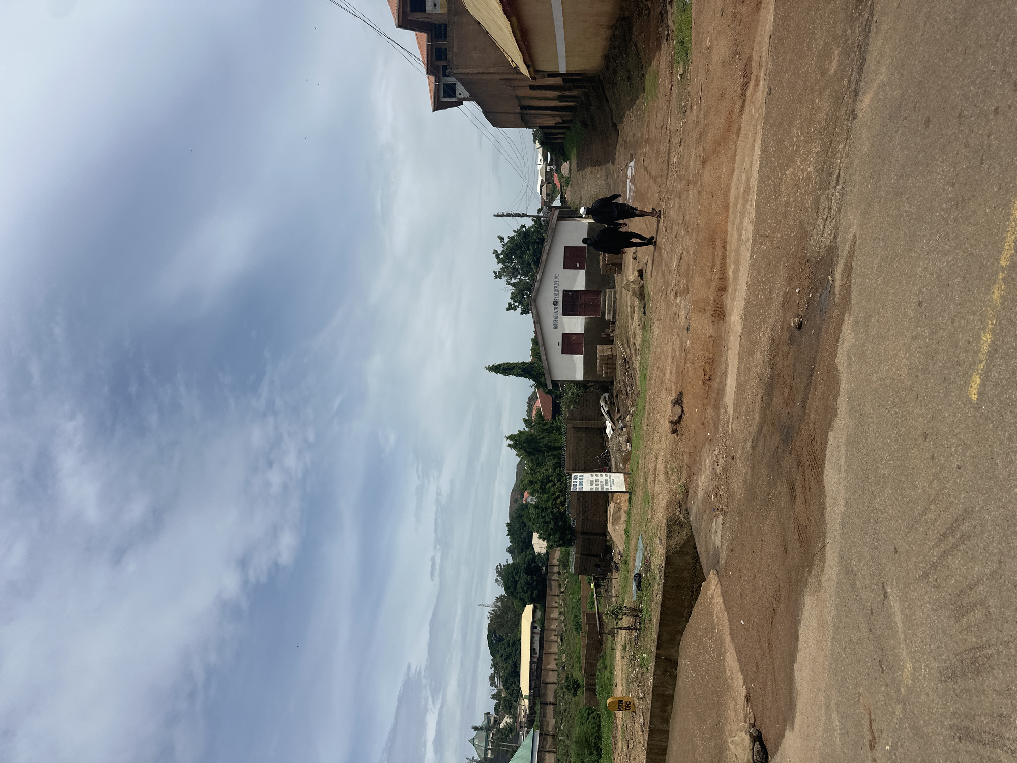 The image depicts a street scene in Jos, Nigeria, under a cloudy sky. In the foreground, a paved road with a yellow line marking transitions to a dirt road. Two individuals, both appearing to be young men, walk away from the viewer down the dirt road. One is wearing a black hoodie and dark pants, while the other wears a black hoodie and shorts with a white cap.
To the left of the dirt road, a low-lying structure with a white facade and red doors is visible, bearing the sign "HOUSE OF FAITH CHURCH INT. INC." Brickwork is stacked to the left of the church entrance. Further into the background, a long, walled property with a yellow roof is seen, bordered by a brick wall. Trees and other buildings are scattered across the landscape, suggesting a mixed urban and semi-rural environment.
On the right side of the dirt road, a tall, multi-story building with a tan facade and a reddish-orange roof is prominent. Adjacent to it, another building with a tan facade and a yellow roofline is partially visible. The overall impression is of a developing area, with elements of construction and established buildings coexisting. The ground is uneven, with patches of dirt, gravel, and some puddles of water. The environment appears to be a typical neighborhood street in Jos.