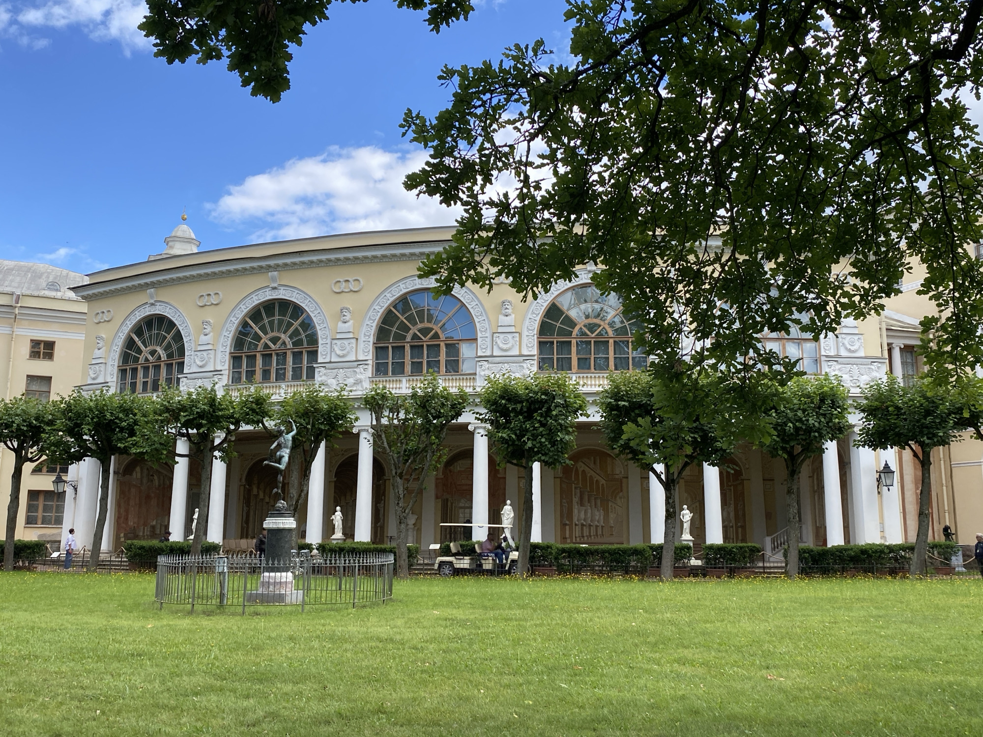 The image depicts the colonnade of the Pavlovsk Palace in Pavlovsk, Russia, on a bright, cloudy day. The facade of the palace is a pale yellow with white architectural details. Large arched windows line the upper level. In front of the colonnade, a row of evenly spaced, manicured trees stands along a low hedge. White columns support the upper story, and several white statues are visible within the arched recesses.
In the foreground, a neatly mown green lawn stretches towards the palace. A dark metal fence encloses a bronze statue of a figure in a dynamic pose, standing on a pedestal. Two figures, possibly tourists, are visible near the statue. Further to the right, a golf cart carrying two individuals is parked near the hedge. Another person stands to the far right of the frame. Lush green foliage from an overhanging tree branches frames the top of the image, casting shadows on the scene below. The sky is a vibrant blue with scattered white clouds.