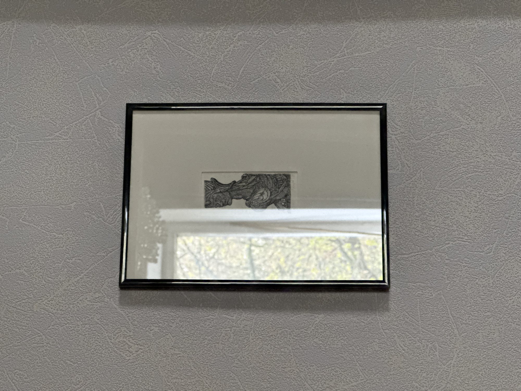 A framed drawing is mounted on a textured, gray wall. The drawing within the frame appears to be a monochromatic sketch of a detailed, complex design. The window behind it reflects the outdoors, showing trees and daylight, which suggests a location in an urban environment. The location appears to be in Moscow, Russia as the windows are identical to those in Russian apartment buildings. The frame is black and reflective.