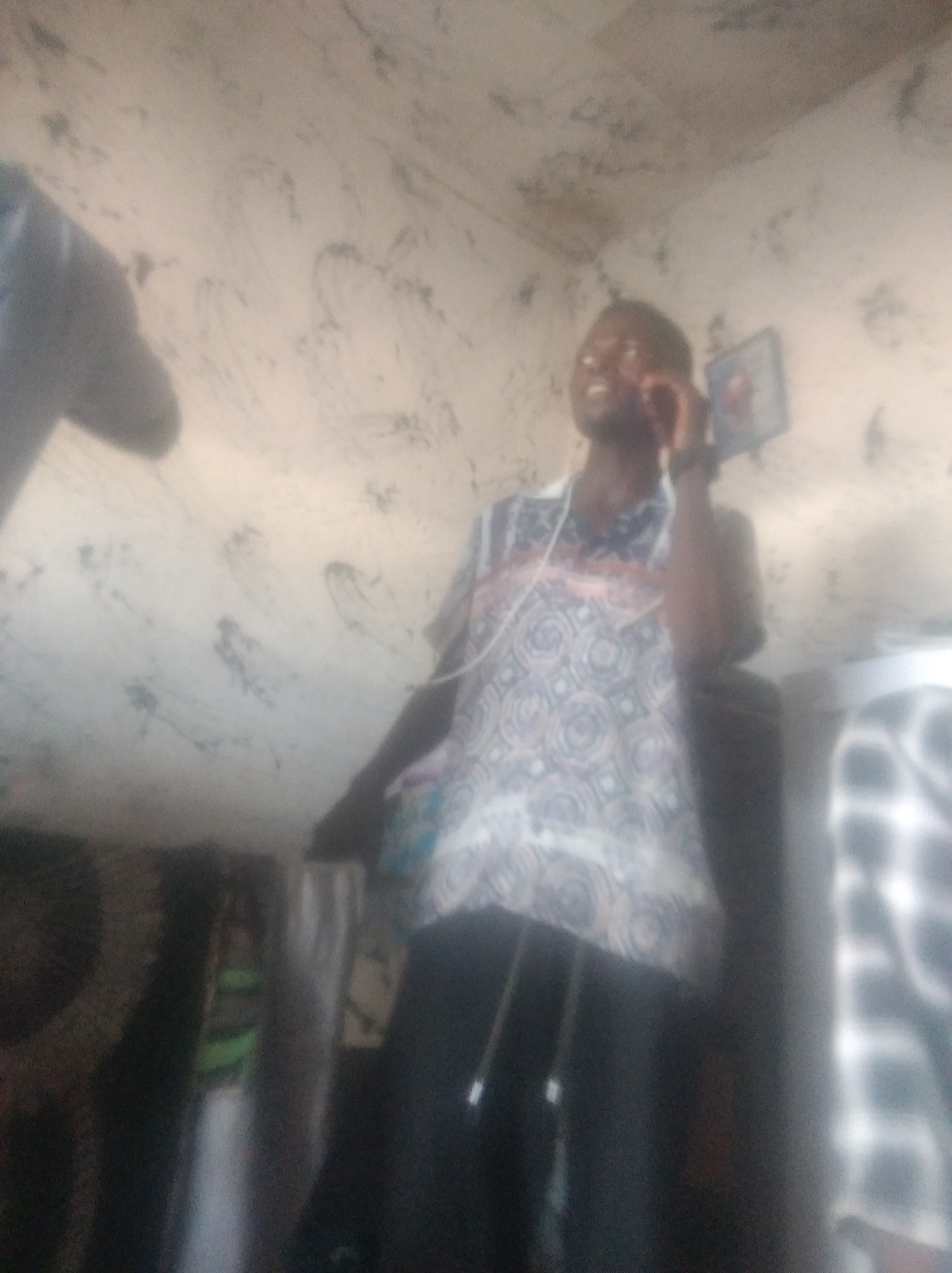 The image depicts a person holding a phone to their ear, seemingly engaged in a conversation. The person is wearing a patterned shirt and dark pants. The background appears to be a room with a textured wall. To the left of the main subject, another person is partially visible, also in dark clothing. On the right side, a portion of another person's checkered shirt is visible. The lighting in the scene is dim, suggesting it might be indoors or late in the day. The overall quality of the image is low, with blurriness obscuring fine details. The context of the activity, such as the nature of the phone call or the setting, is not clearly discernible.