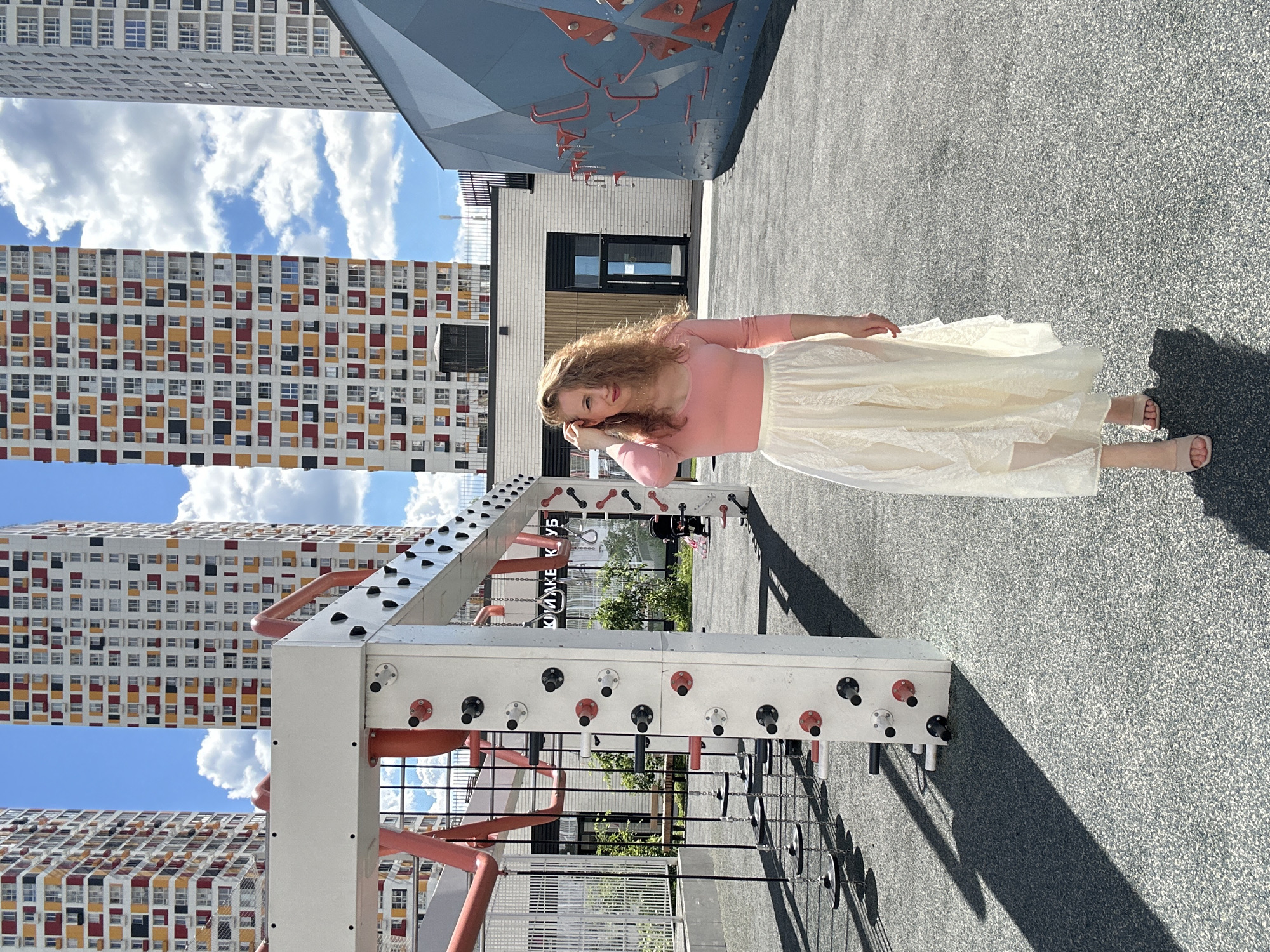 A woman with curly blonde hair stands in an outdoor recreation area in Krasnogorsk, Russia. She is wearing a pink long-sleeved shirt and a long, flowing white lace skirt. She is standing on a grey, speckled surface, likely rubberized playground flooring. Behind her are modern apartment buildings with colorful facades. To her left is a section of an outdoor climbing wall, and to her right is a children's playground structure with climbing elements. The sky is blue with scattered white clouds. The lighting suggests it is daytime, possibly late morning or early afternoon.