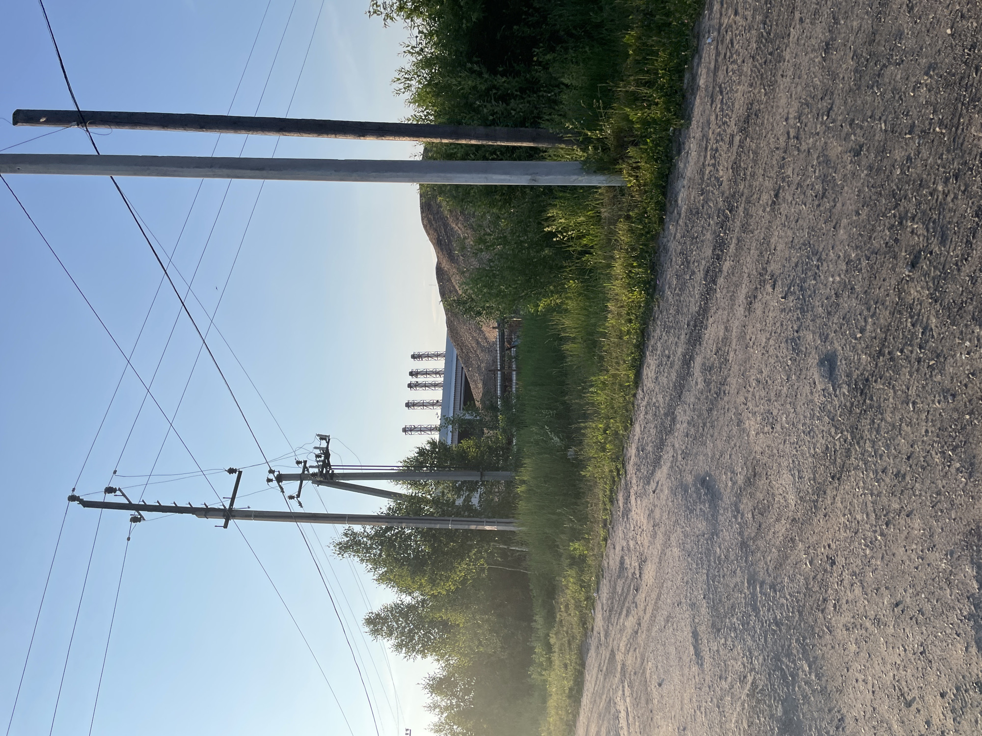 The image depicts a rural scene near the city of Prudy, Russia. A dirt road occupies the foreground, with patches of gravel and tire tracks visible. To the left of the road, lush green grass and vegetation grow, extending towards a line of trees. Beyond the trees, several utility poles with wires are visible against a clear blue sky. In the background, industrial structures with tall chimneys rise above a large pile of what appears to be woodchips or debris. The lighting suggests it is either early morning or late afternoon, with a gentle sunlight casting soft shadows. There are no people or animals present in the image.