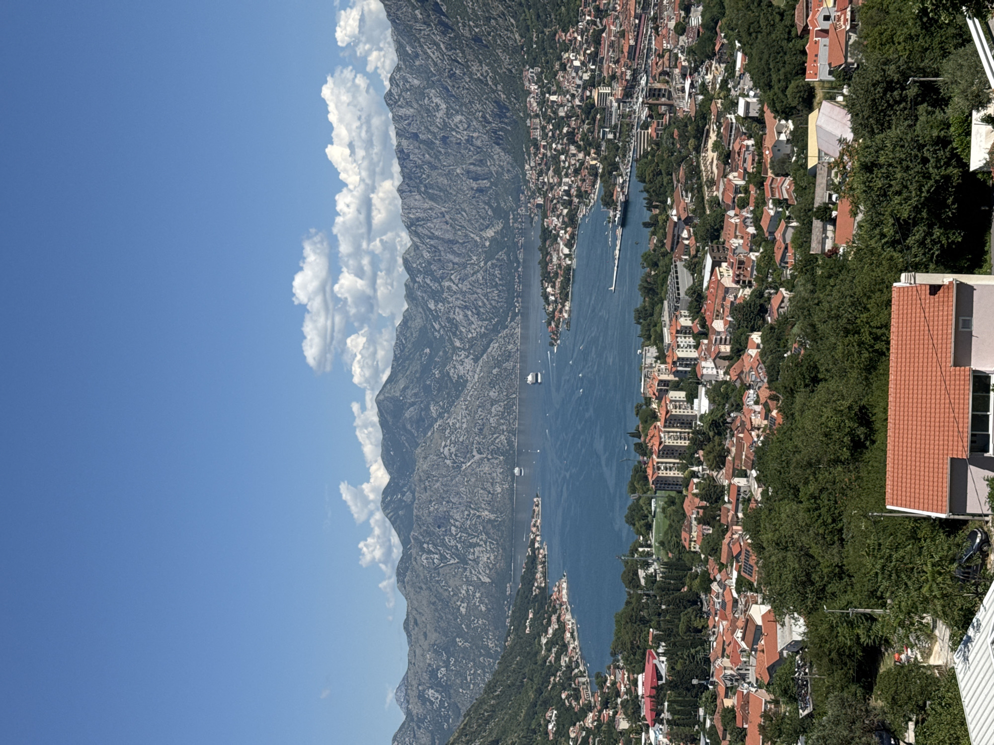 This is a high-angle, long-shot, vertical photograph taken on a sunny day with a clear blue sky and scattered white clouds. The scene depicts a large bay or fjord surrounded by majestic, rugged, gray mountains with sparse vegetation. The water of the bay is a deep blue, with several white cruise ships and smaller boats visible on its surface.
Along the shoreline of the bay, a dense, urban settlement stretches out. The architecture is predominantly composed of buildings with red-tiled roofs, giving the cityscape a cohesive visual theme. Some of these buildings are mid-rise apartments and commercial structures, while others appear to be smaller residential homes. Green trees are interspersed throughout the urban area and also cover the lower slopes of the surrounding mountains, creating a stark contrast between the built environment and the natural landscape.
In the foreground, more of the red-roofed buildings are visible, some with solar panels on their roofs. A few vehicles are parked on what appears to be a paved area or road. The overall impression is of a vibrant coastal town nestled within a dramatic natural setting.