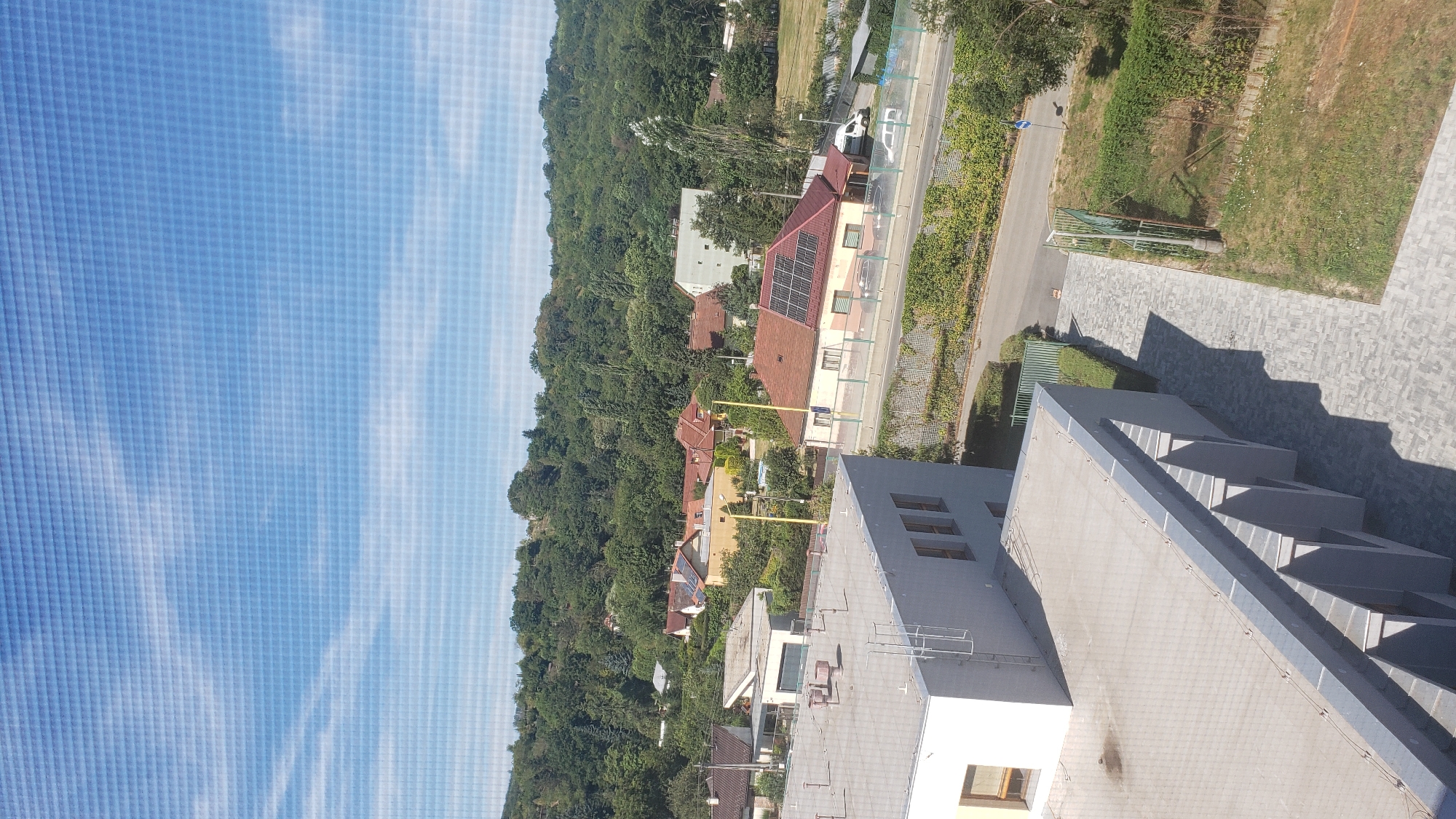 This is an aerial, outdoor, daytime photograph showing a residential area with trees and buildings. The foreground is dominated by the roof of a modern building with a grey, flat roof and sections of grey siding. There are several windows visible on the side of this building. To the right of this building, a grey paved walkway leads past a green metal fence and a grassy area with some sparse vegetation.
In the middle ground, a street is visible with a concrete barrier and some parked vehicles, including a white van and a white car. Adjacent to the street is a building with a red-tiled roof covered in solar panels. Further back, several other houses with red or orange tiled roofs are scattered amongst dense green trees. The background features a hillside covered in a thick canopy of trees. The sky above is blue with scattered white clouds.
This image appears to be taken from an elevated position, likely a building, looking out over a neighborhood that is a mix of residential houses and some larger structures, with a significant amount of greenery. The presence of solar panels on one of the houses suggests a focus on renewable energy. The street and the presence of multiple vehicles indicate a developed residential area.
Based on the visual cues, specifically the architectural style of the houses, the type of vegetation, and the overall landscape, this scene is consistent with residential neighborhoods found in Prešov, Slovakia, particularly in areas where the urban environment meets the surrounding hillsides. However, without specific landmarks or signage, a precise location within Prešov cannot be determined.