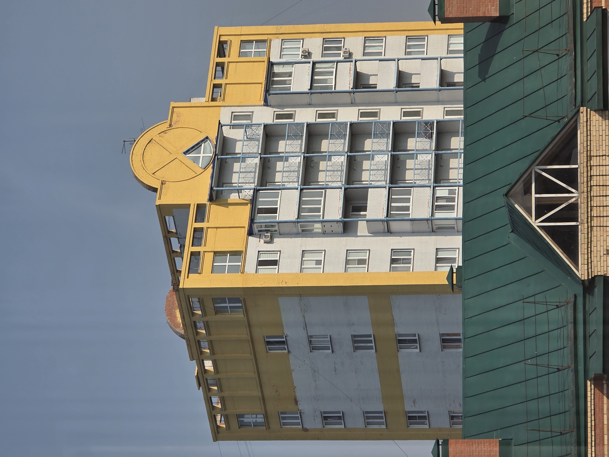 A tall, modern residential building with a yellow and gray facade stands against a clear, pale blue sky. The building features multiple stories with evenly spaced windows and balconies, many of which are enclosed with light blue railings. The top of the building is adorned with a distinctive yellow decorative element, reminiscent of a sun or a stylized "X" shape, with a triangular window set within it. A reddish-brown dome is visible on the rooftop. In the foreground, a green, metal-pitched roof with visible seams and some supporting structures partially obscures the lower portion of the residential building. The scene suggests an urban environment, possibly in Omsk, Russia, given the architectural style. No people are visible in the image.