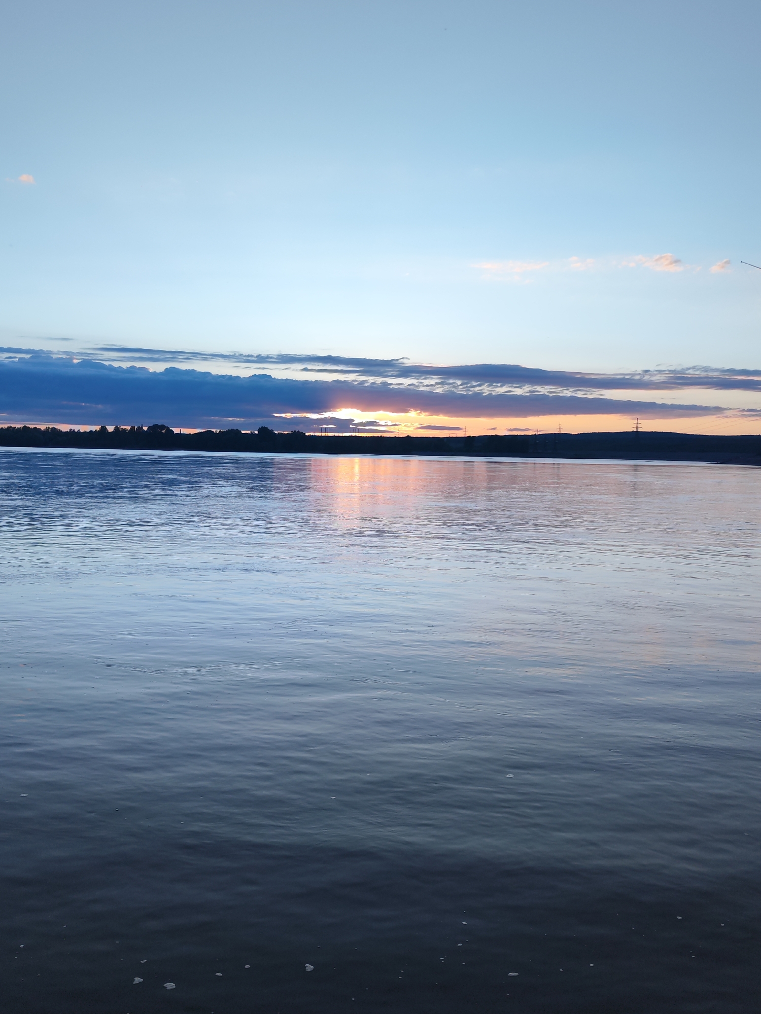 The image presents a tranquil sunset scene over a wide body of water, likely a river or reservoir in Chaikovsky, Russia. The sky dominates the upper portion of the frame, transitioning from a clear, pale blue at the top to layered clouds in various shades of grey and deep blue across the mid-section. Closer to the horizon, the sky bursts with warm hues of orange, yellow, and soft pink as the sun descends.
The sun is partially obscured by a dark, silhouetted landmass on the horizon and the surrounding clouds, creating a vibrant glow that reflects on the water's surface. This distant landmass appears as a low bank or hills, with subtle indications of vertical structures, possibly utility poles or power lines, particularly on the right side.
The expansive water in the foreground is calm, displaying gentle ripples and reflecting the soft blues and warmer sunset tones of the sky. The reflection of the sun creates a bright, shimmering path across the water. The time of day is clearly dusk or early evening, characterized by the low sun and the rich, transitioning colors of the sky. The scene conveys a sense of peaceful natural beauty. No people, discernible activities, or visible text are present in the image.