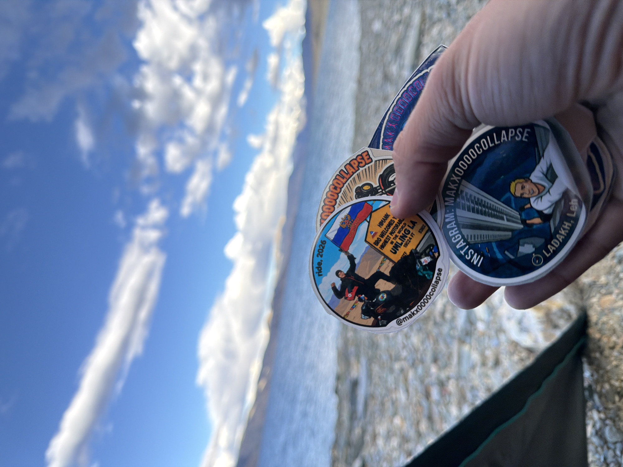 A person's hand holds several round stickers in the foreground, with a blurred background of a body of water and mountains. The stickers depict a person on a motorcycle, a Russian flag, and an urban landscape. One sticker contains the text "INSTAGRAM: MAXXOOOCOLLAPSE LADAKH, LEH". The environment appears to be rocky and barren, consistent with the high-altitude desert landscape of Ladakh. The weather is partly cloudy with a bright blue sky.