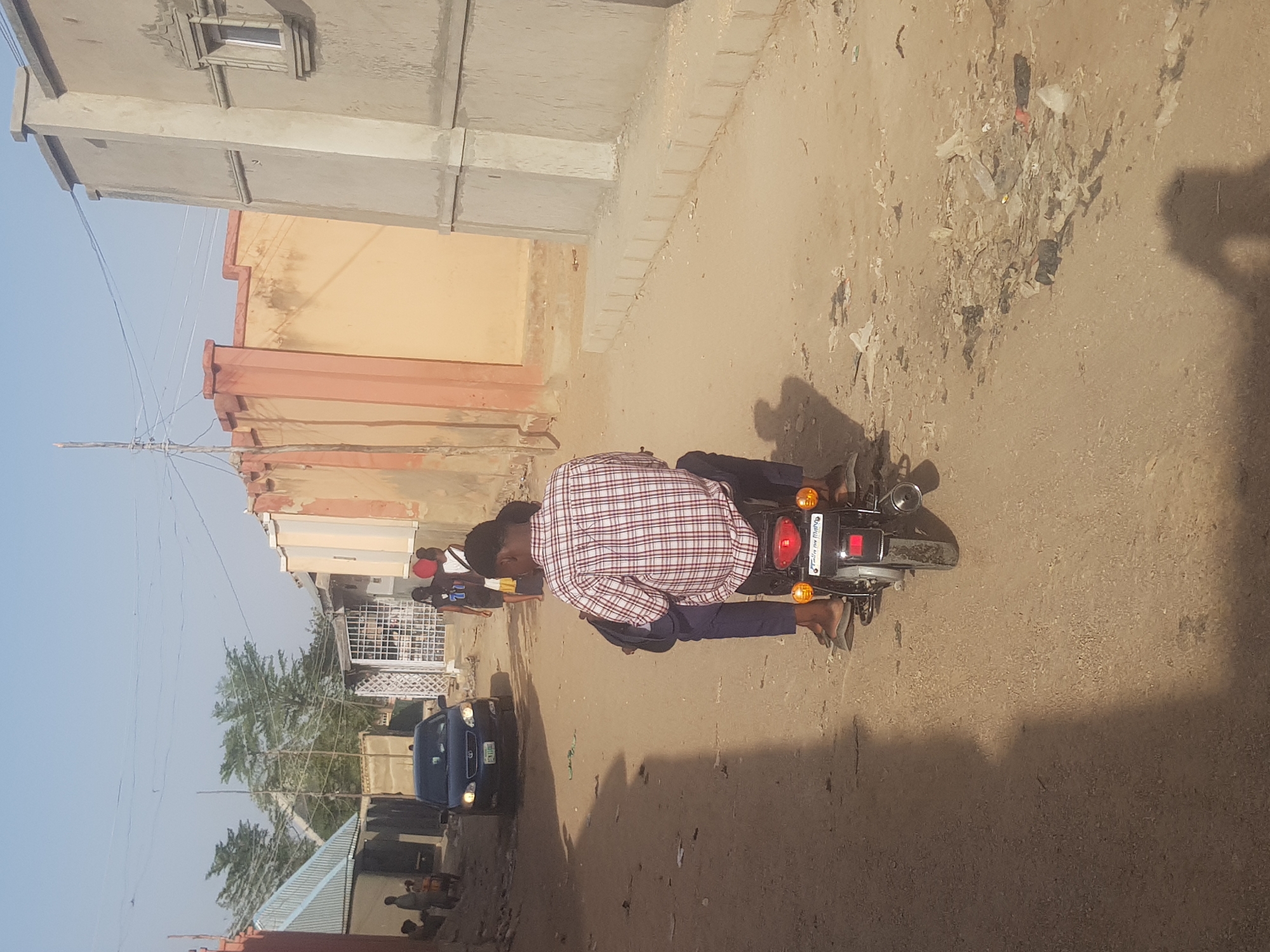 A street scene in Dutsinma A, Nigeria, features a person on a motorcycle, oriented perpendicularly to the road. The individual, wearing a plaid shirt and dark pants, is seated on the motorcycle. Nearby, a blue car is parked. Other people are visible in the distance. The road is unpaved, and there's debris scattered on the ground. Buildings line one side of the road. The sky is clear and light blue.