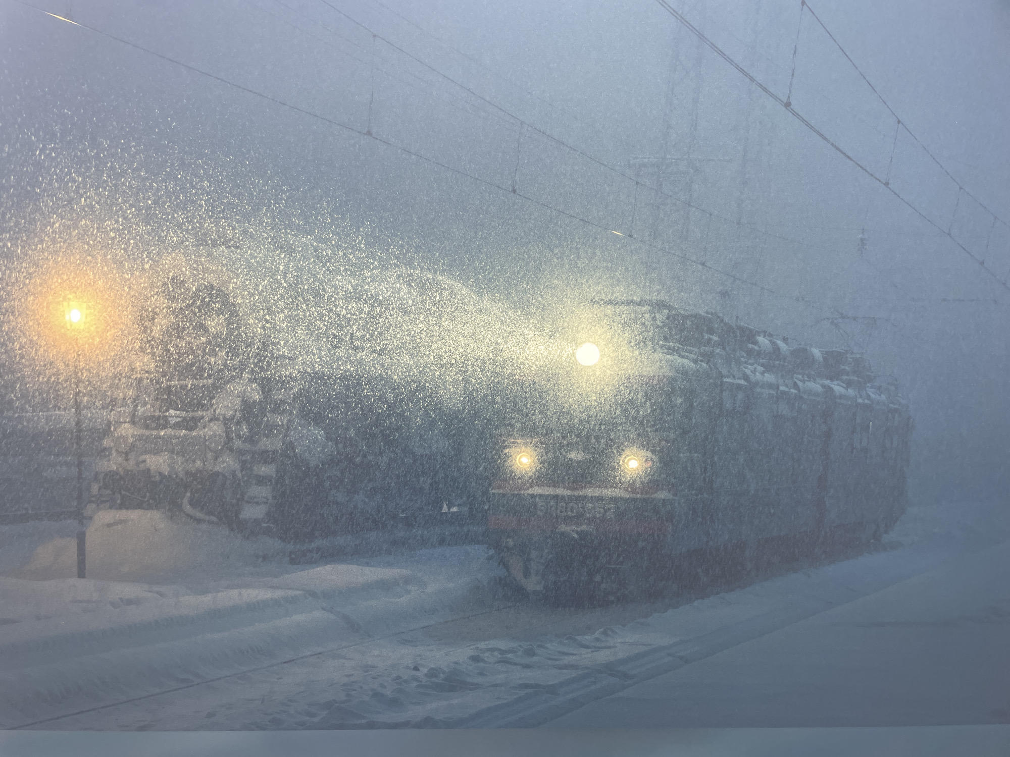A train is moving through heavy snowfall in Sochi, Russia. The train's headlights are visible, and there is a light post illuminated by a golden glow on the left. Tracks are covered in snow. Overhead wires are visible. A second train may be present on a track to the left.