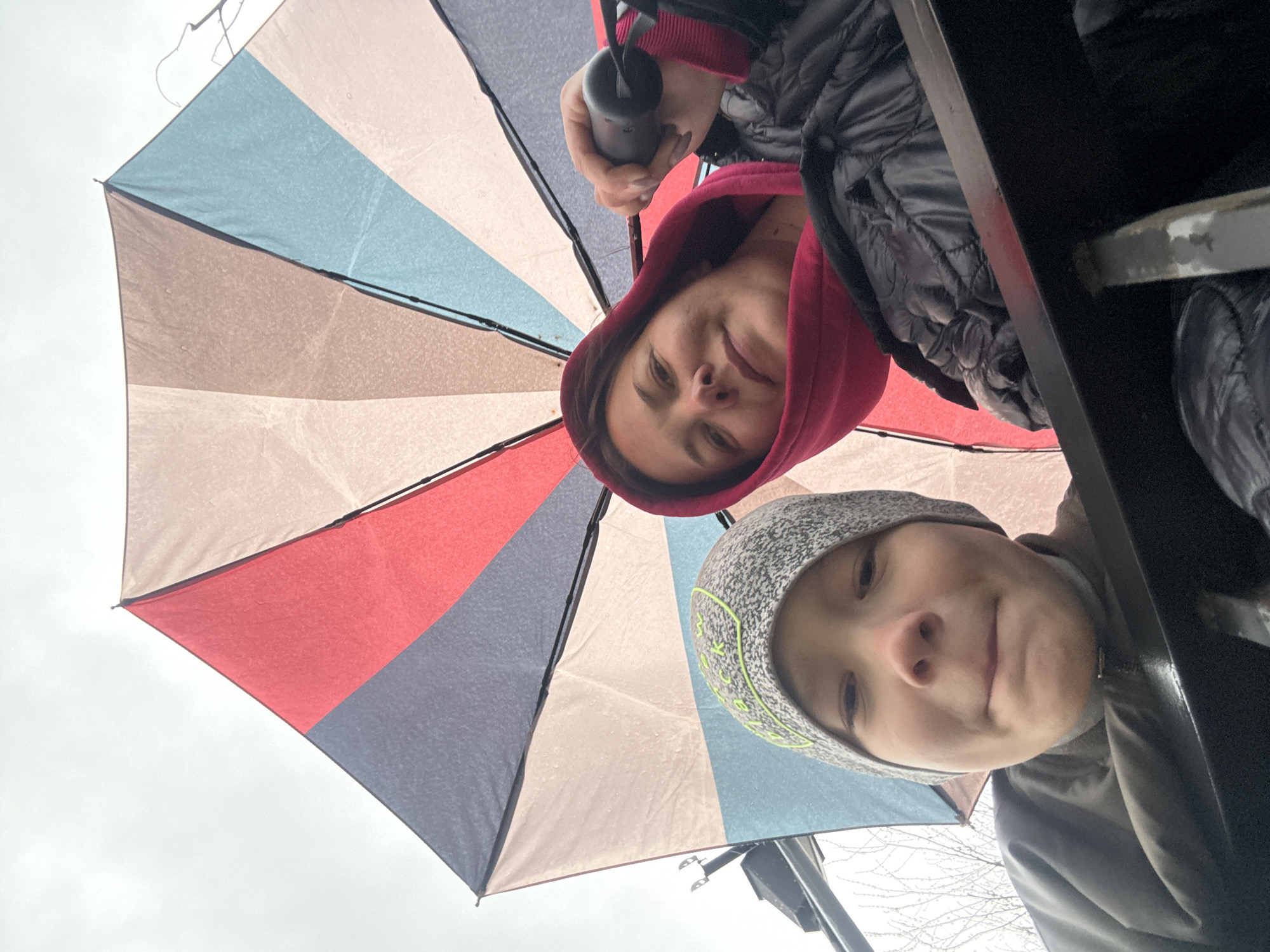 A low-angle shot captures a woman in a red hooded sweatshirt and a black puffer jacket holding a multicolored umbrella over herself and a child wearing a gray knit hat. The sky is overcast and the umbrella canopy is divided into sections of light blue, brown, beige, red, and gray. Metal structures are partially visible on the right. Based on the location, this may have occurred in Dmitrov, Russia.