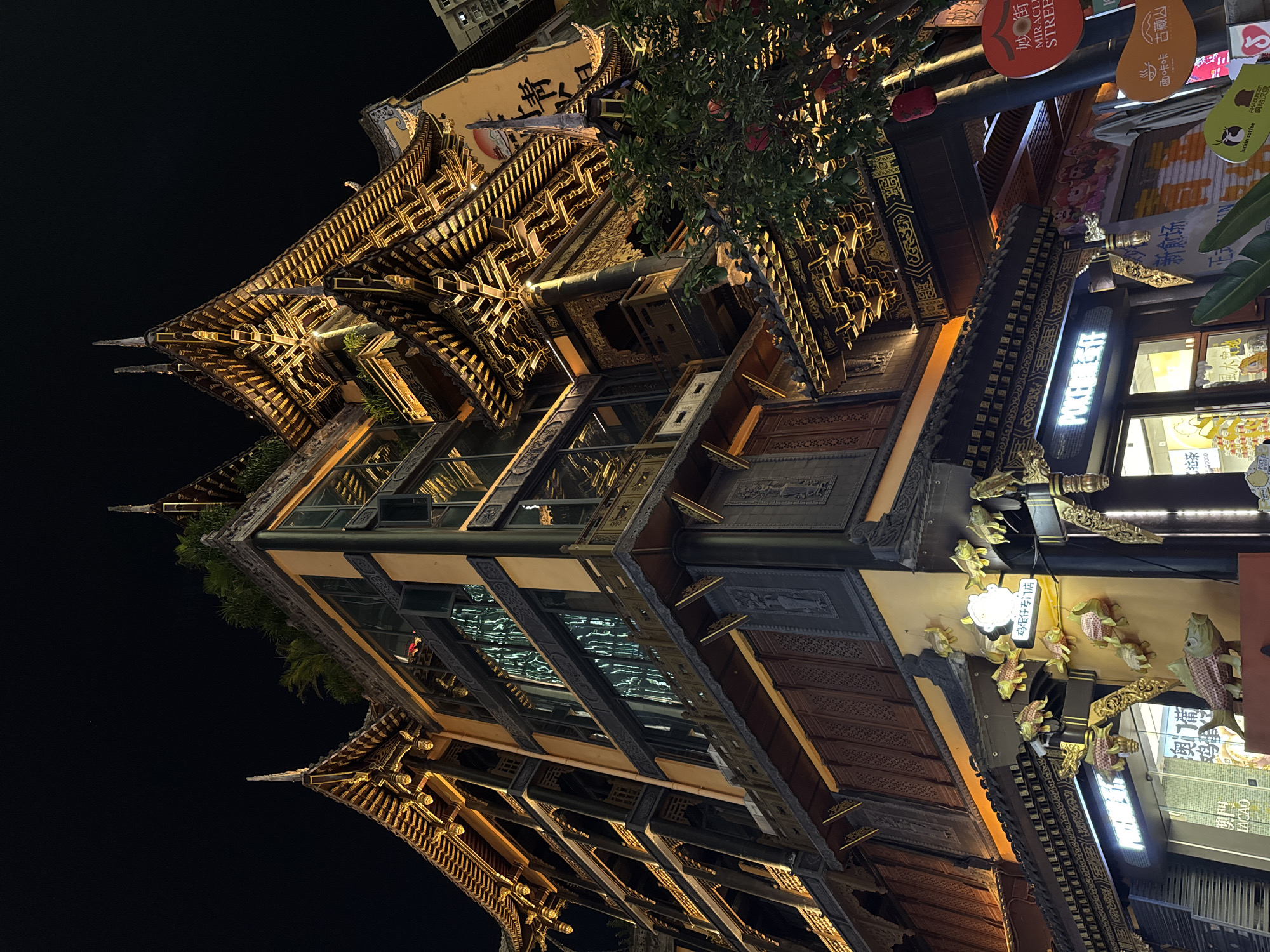 The image depicts a multi-story building at night, characterized by traditional Chinese architectural elements. The building's exterior features ornate, tiered roofs with exposed wooden beams and decorative eaves, illuminated by warm lighting. The facade is a mix of dark wood, yellow ochre walls, and intricate carvings. Large glass windows are visible on multiple levels, reflecting the surrounding lights.
On the lower levels, storefronts are visible, with neon signs and illuminated displays. One sign reads "POKER" in English, and another appears to be in Chinese characters. Decorative elements, such as stylized golden fish and relief carvings, adorn the exterior walls and awnings. A tree with red fruit and lanterns is situated to the right of the building. The sky is dark, indicating nighttime.
The scene is located on a street in Yuzhong District, Chongqing, China, as indicated by a street sign that reads "Miracle Street" and other Chinese text, likely referencing a specific street or area. The architecture suggests a commercial or entertainment district. No people are clearly visible in the image.