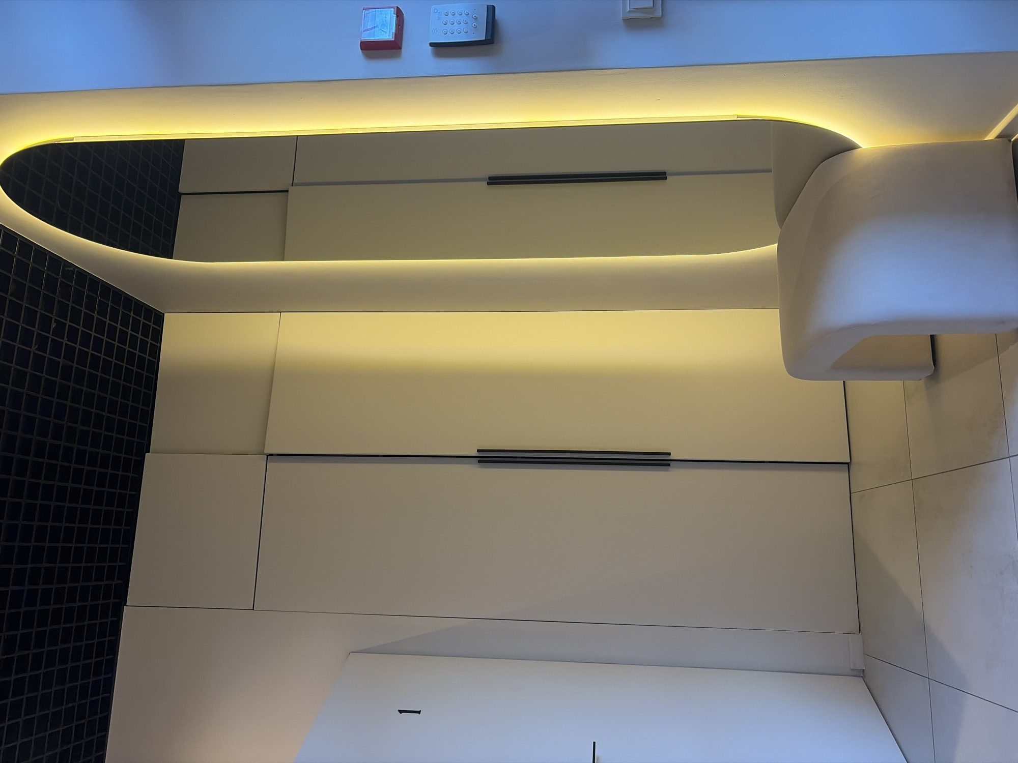 A photograph shows a modern interior space. Two rows of cabinetry with horizontal handles are the primary focus, illuminated by a warm, indirect light source above. The left side of the image features a dark, small-tiled wall, contrasting with the white cabinetry. On the right, a portion of a white chair is visible, along with light-colored tiles on the wall. Near the top of the frame, a red fire alarm and a grey security panel are mounted on the wall. The cabinets are white and appear to be located within a bathroom. The reflection in the mirror gives the appearance of a rounded shape. Without specific context, such as signage or distinctive landmarks, pinpointing the exact location within Saint Petersburg, Russia, is not possible based on the image alone.