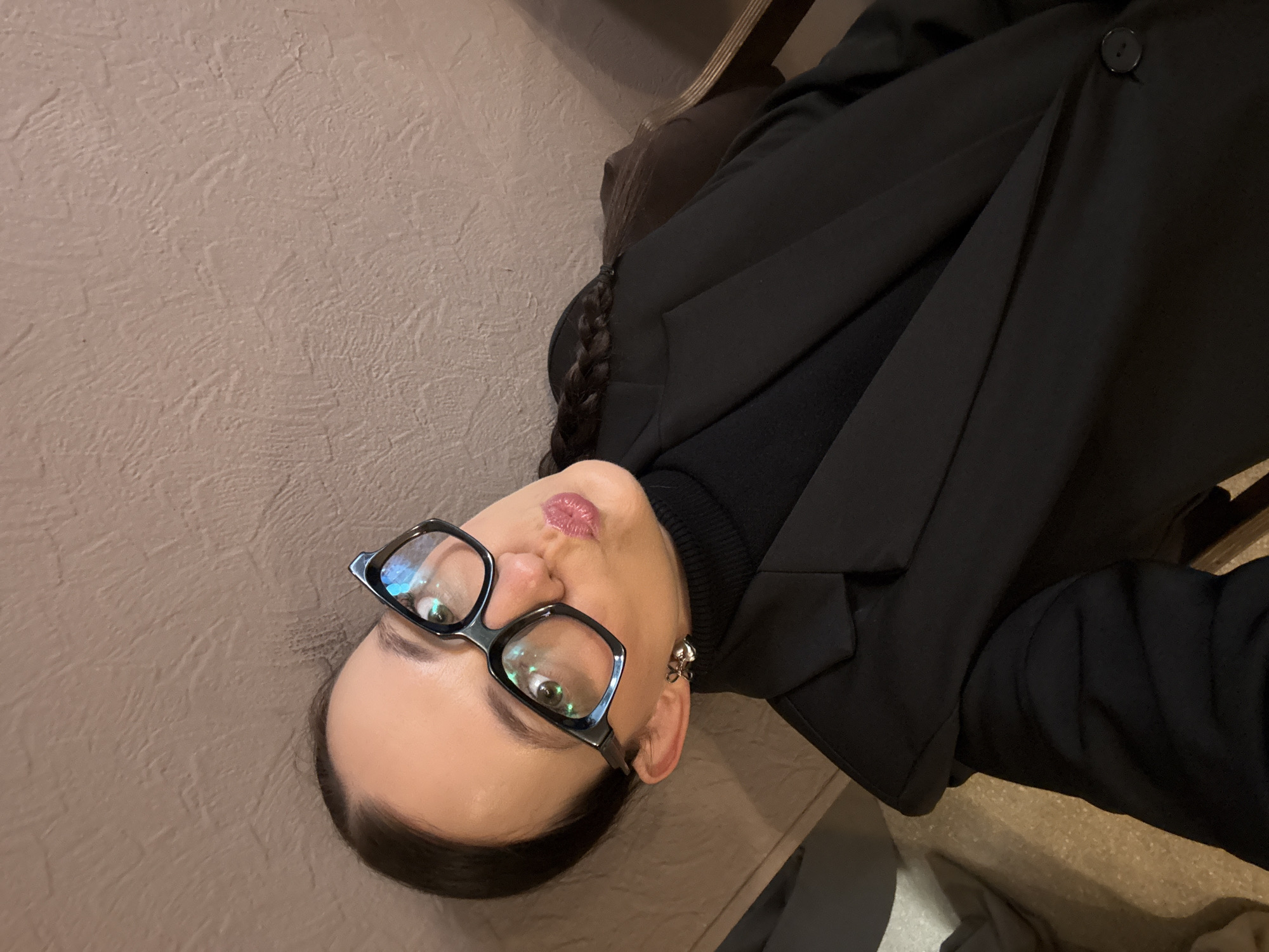 A woman with dark hair styled in a braid and wearing glasses, a black turtleneck, and a black blazer is taking a selfie. Her lips are pursed as if she is blowing a kiss. She is wearing a silver earring in the shape of a bow on her left ear. The background is a textured brown wall. The lighting is dim, suggesting an indoor setting, possibly a restaurant or cafe. There is no indication of the location being in the city of Chaikovsky, Russia.