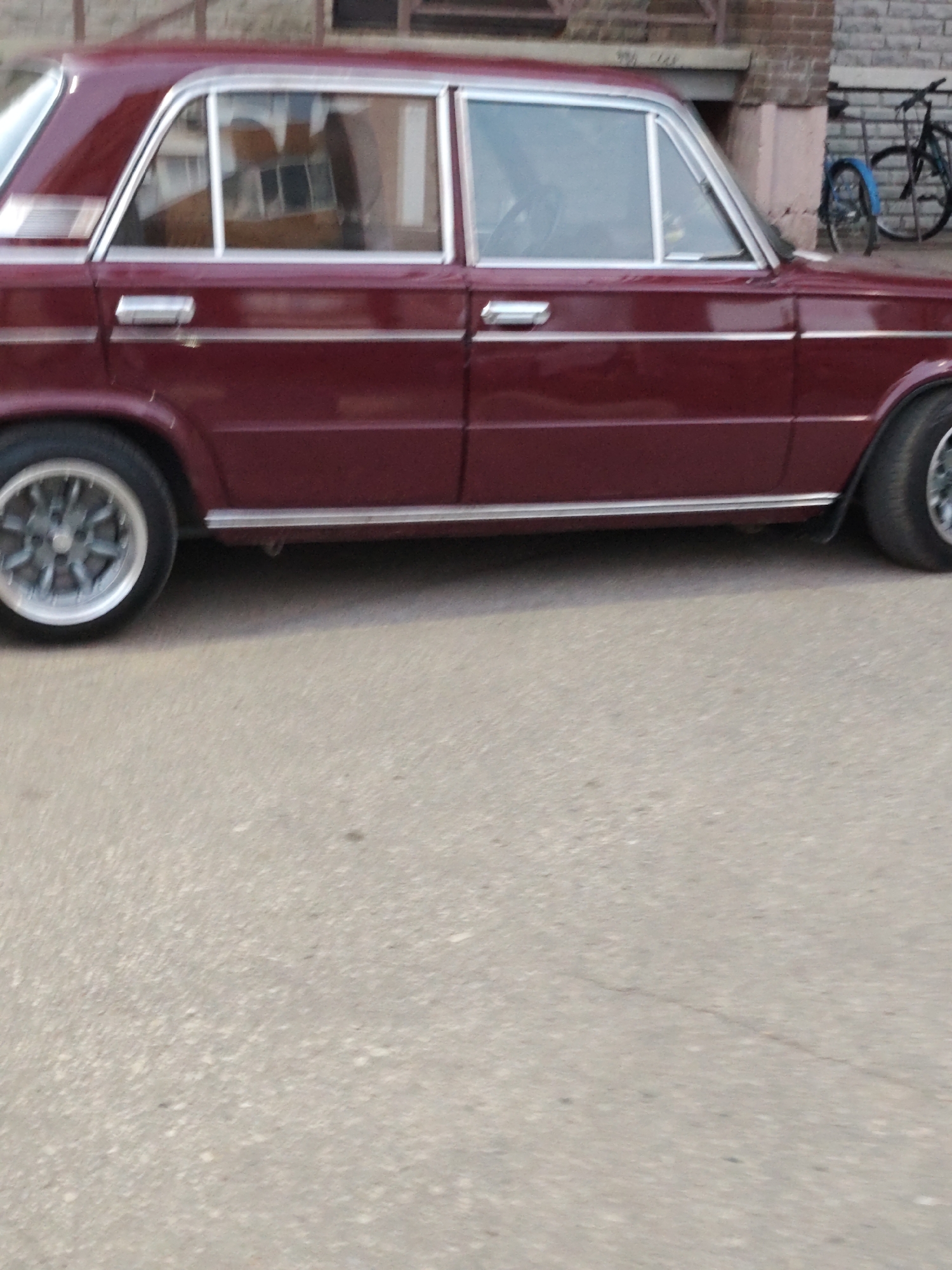 The image shows a side view of a maroon sedan, likely a Lada, parked on an asphalt surface. The car has chrome trim along the bottom of the doors and silver multi-spoke wheels. In the background, a brick building with windows is visible, and to the right, a bicycle with a blue fender is leaning against a fence. The overall scene appears to be an urban environment, possibly in Syktyvkar, Russia, based on the request. The lighting suggests it is daytime.
