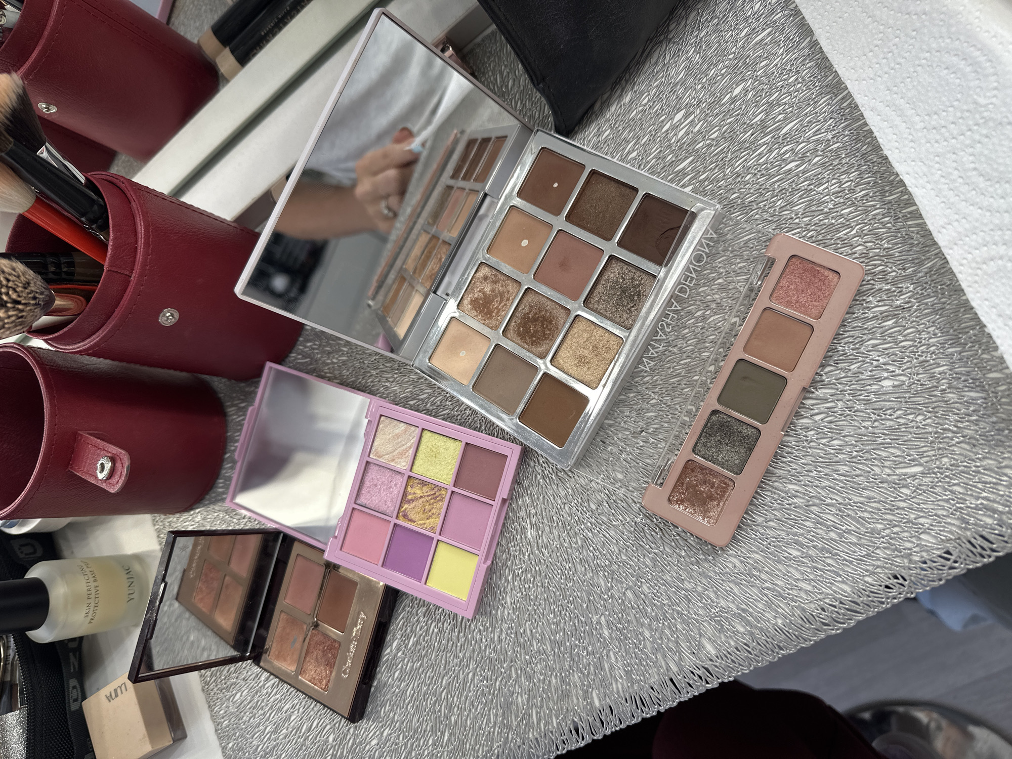 This media file is a close-up, overhead shot of various makeup products laid out on a silver, textured surface. The subjects are makeup palettes and brushes. In the background, there are red leather brush holders and a black makeup bag. A hand with a ring is visible in the reflection of one of the palettes, suggesting the photo was taken by someone holding the palette.
The objects visible include:
- A large, rectangular eyeshadow palette with twelve neutral-toned shades, open and angled to show the individual pans.
- A smaller, square eyeshadow palette with nine pastel shades, including pinks, purples, and yellows, also open.
- A small, rectangular eyeshadow palette with three shades, including a rose-gold shimmer, a light brown, and a dark olive green.
- A partially visible makeup palette with four shades in warm, coppery tones, reflected in a mirror.
- A bottle of skincare product with text that includes "YUNJAC" and "SKIN PERFECTIVE PROTECTIVE BASE."
- Makeup brushes stored in red cylindrical holders.
- A black makeup bag or pouch.
- A white paper towel in the upper right corner.
The activity taking place is the arrangement and presentation of makeup items, likely for a review, flat lay, or organizational display. There are no specific time of day or weather cues. The setting appears to be a tabletop or vanity.
There is some visible text on the bottle of skincare product, "YUNJAC" and "SKIN PERFECTIVE PROTECTIVE BASE." The word "YUNJAC" is also reflected in the mirror of the palette in the center of the image. The brand "Charlotte Tilbury" is visible on the partially open palette at the bottom.