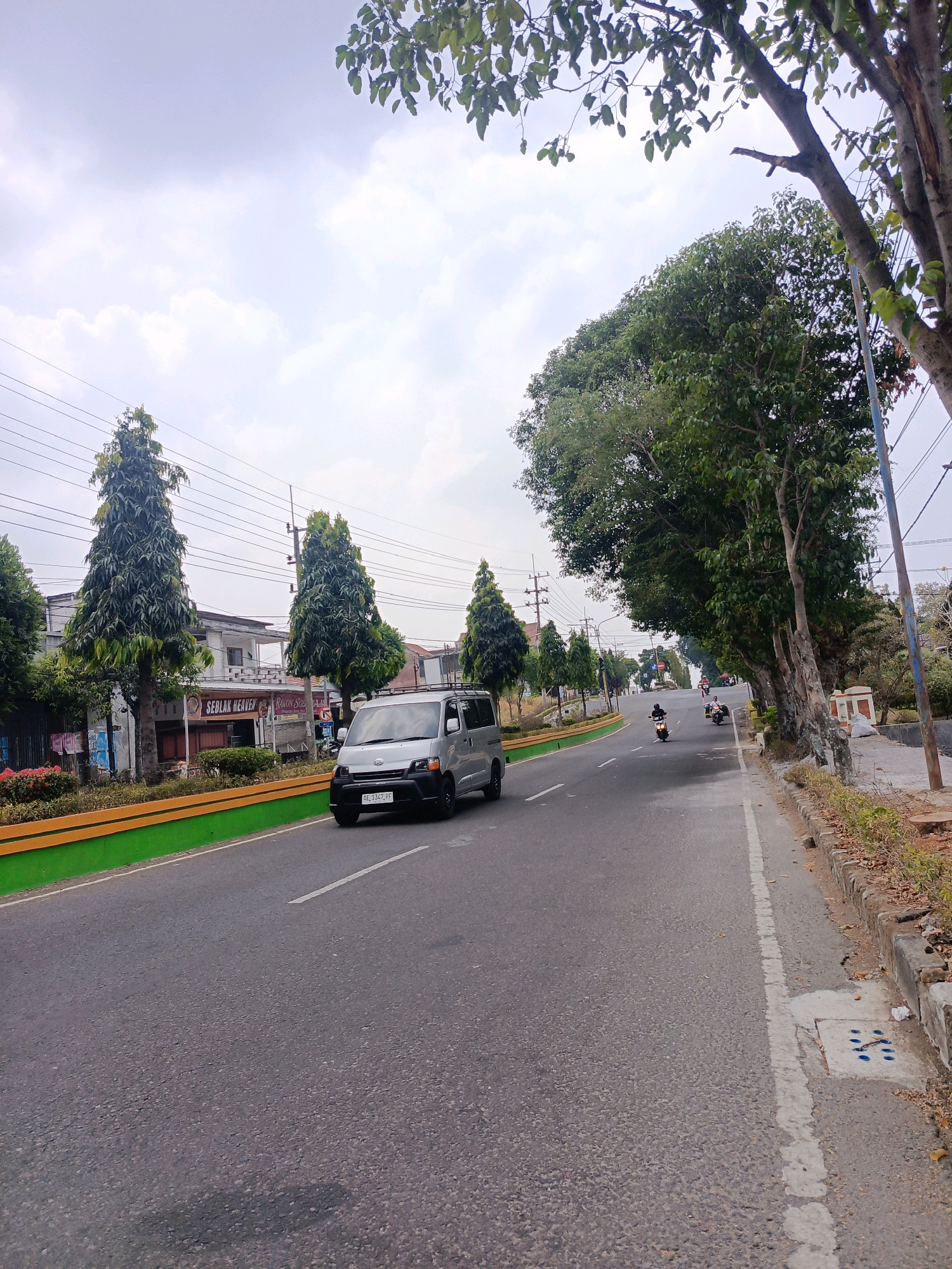 The image depicts a street scene in Sukowinangun, Indonesia, under an overcast sky. A silver van is traveling on the asphalt road, which is lined with trees and vegetation. On the left side of the road, there are buildings, including a shop with a sign that reads "SEBLAK HEAVEN." Several tall, slender trees are planted along the sidewalk. On the right side of the road, a sidewalk is visible with a concrete curb and some fallen leaves. The road also shows lane markings and a white line delineating the edge of the lane. Further down the road, several motorcycles are seen in motion. The overall impression is of a moderately busy street in a semi-urban environment.