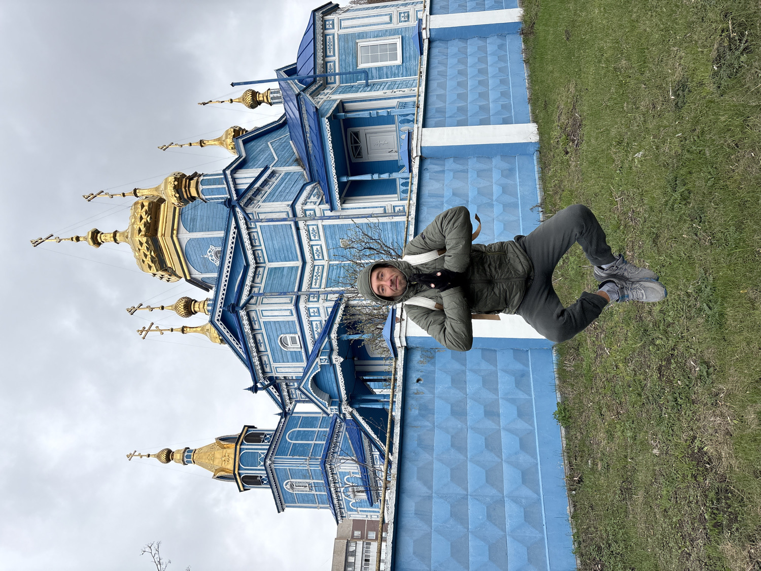 The image features a man standing in front of a striking blue and gold Russian Orthodox church under an overcast sky. The man, positioned slightly to the right of the frame, is wearing a dark olive-green hooded jacket, dark grey pants, and light grey athletic shoes. A light-colored backpack is visible on his shoulders, and he looks directly at the camera with a neutral expression.
Behind the man is a patch of green grass, beyond which stands a blue, textured wall or fence section. The main subject in the background is the church, which boasts an ornate wooden construction painted in a vibrant blue with white decorative trim. It is adorned with multiple golden onion domes, each topped with a cross. The intricate details of the wooden architecture, including window frames and decorative elements, are prominent. The sky is uniformly grey and cloudy, indicating an overcast day. No discernible text is visible in the image. The setting appears to be outdoors, likely a church ground or a public area adjacent to the religious building, consistent with a location such as Ulyanovsk, Russia.